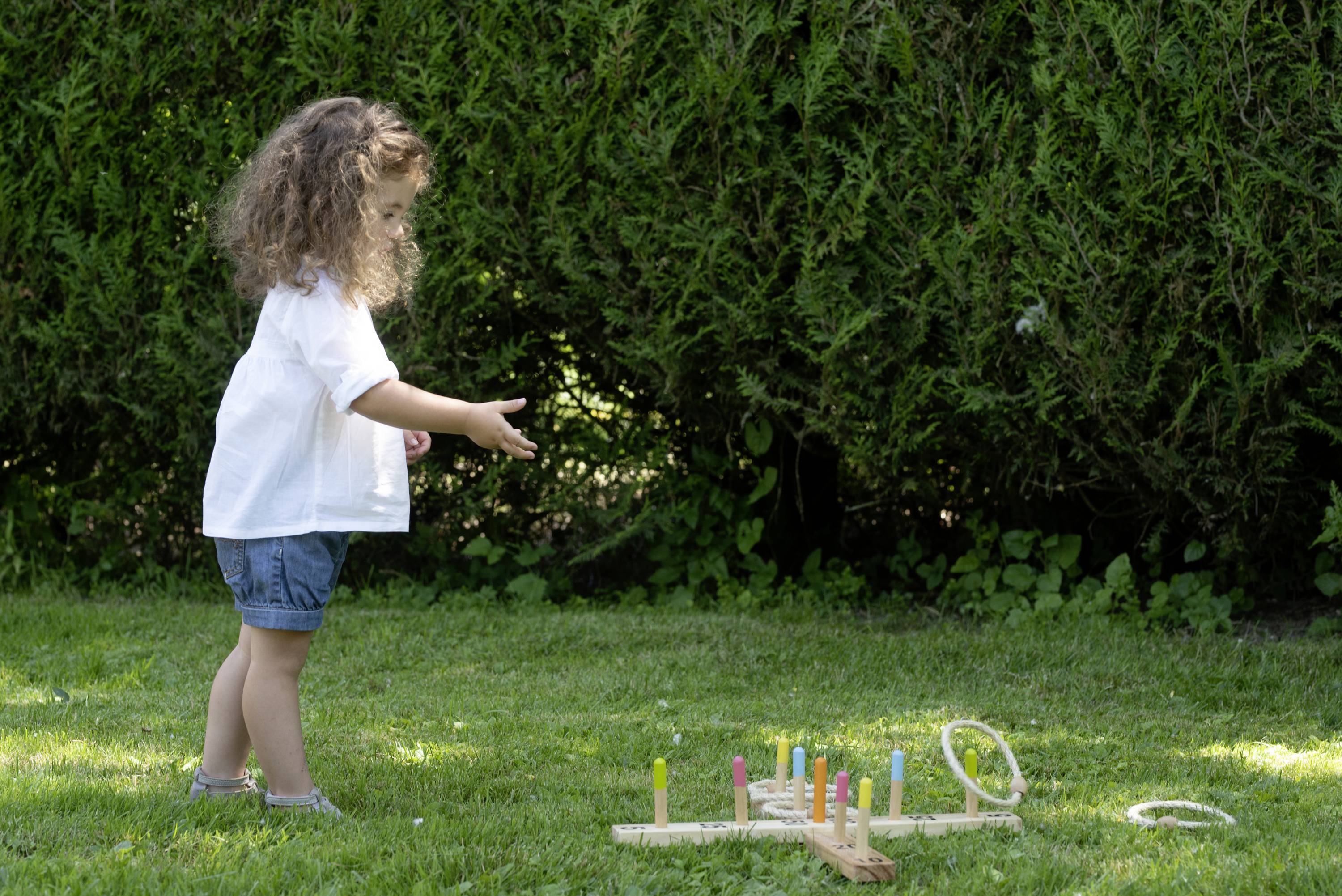 A child with curly hair is playing a ring toss game on the lawn in front of a backdrop of green bushes.