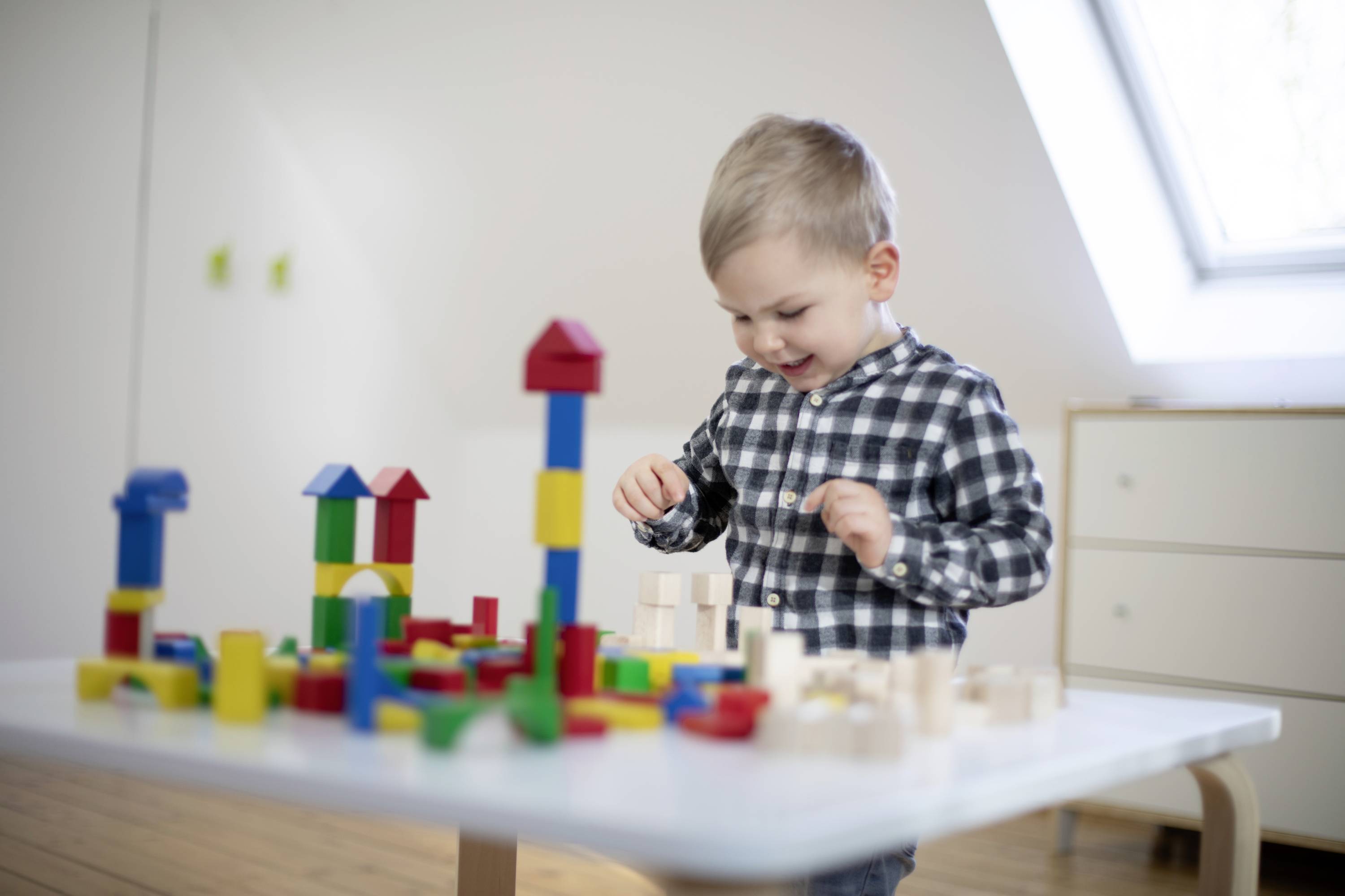 A small child is happily playing with colourful wooden building blocks at a table in a bright room.