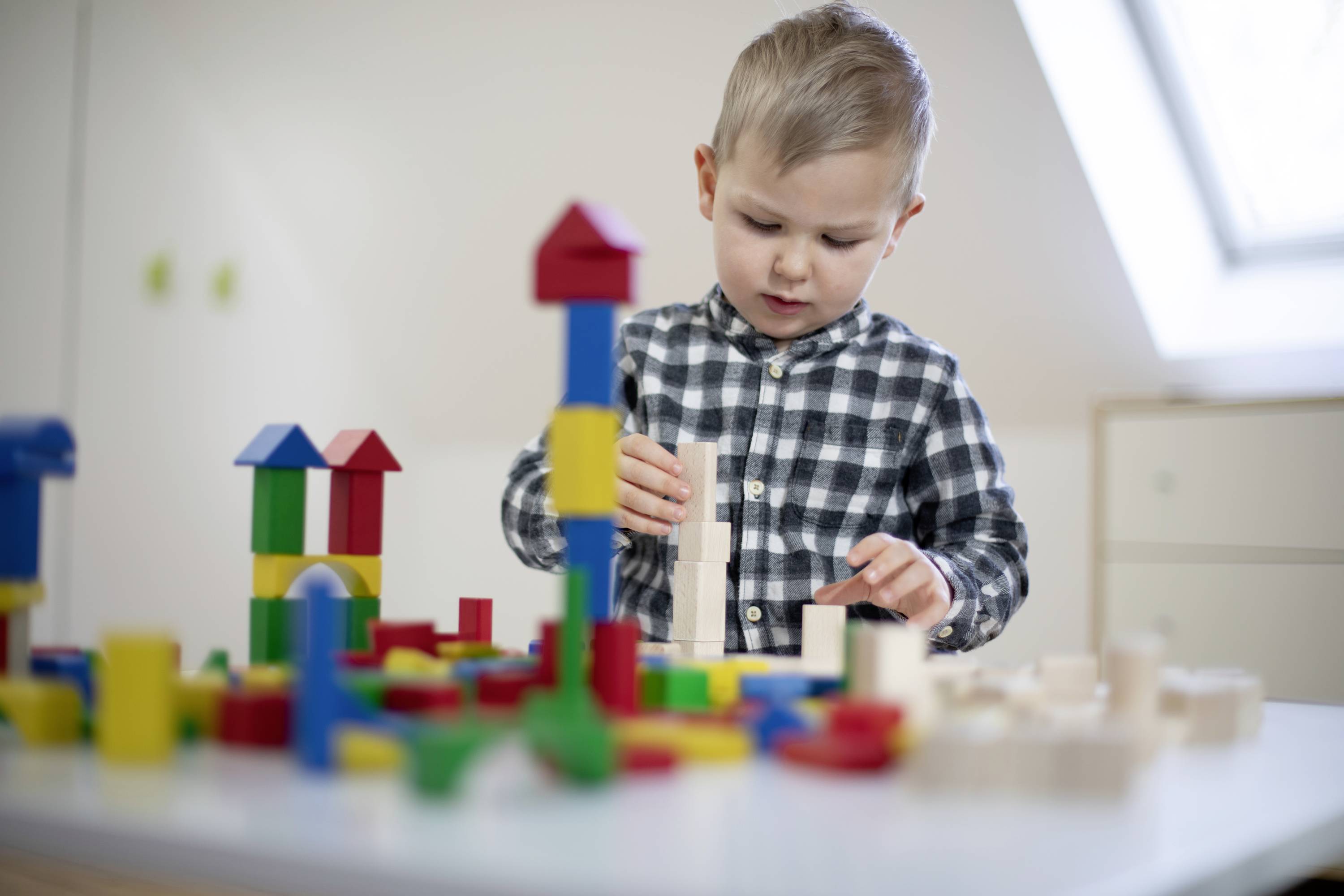 A child plays intently with colourful wooden building blocks on a table, stacking and constructing towers in a bright room.