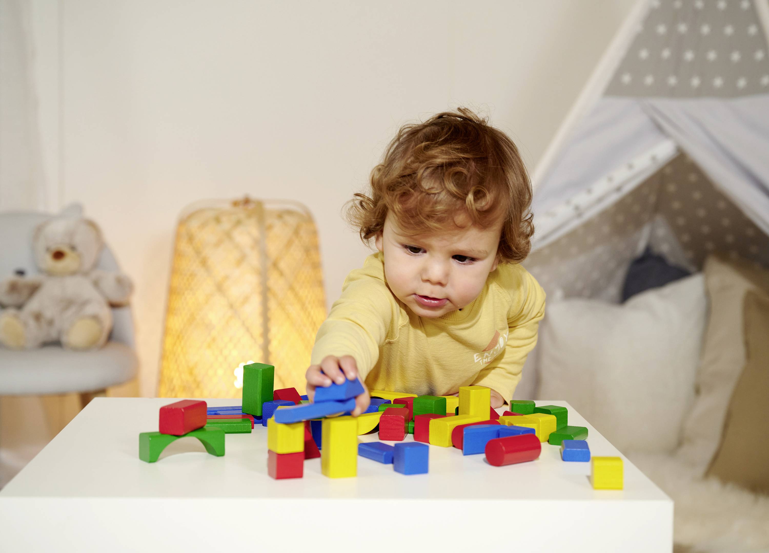 A toddler is playing with colourful building blocks on a table in a cosy room. In the background, a tent and a teddy bear.