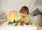 A toddler is playing with colourful building blocks on a table in a cosy room. In the background, a tent and a teddy bear.