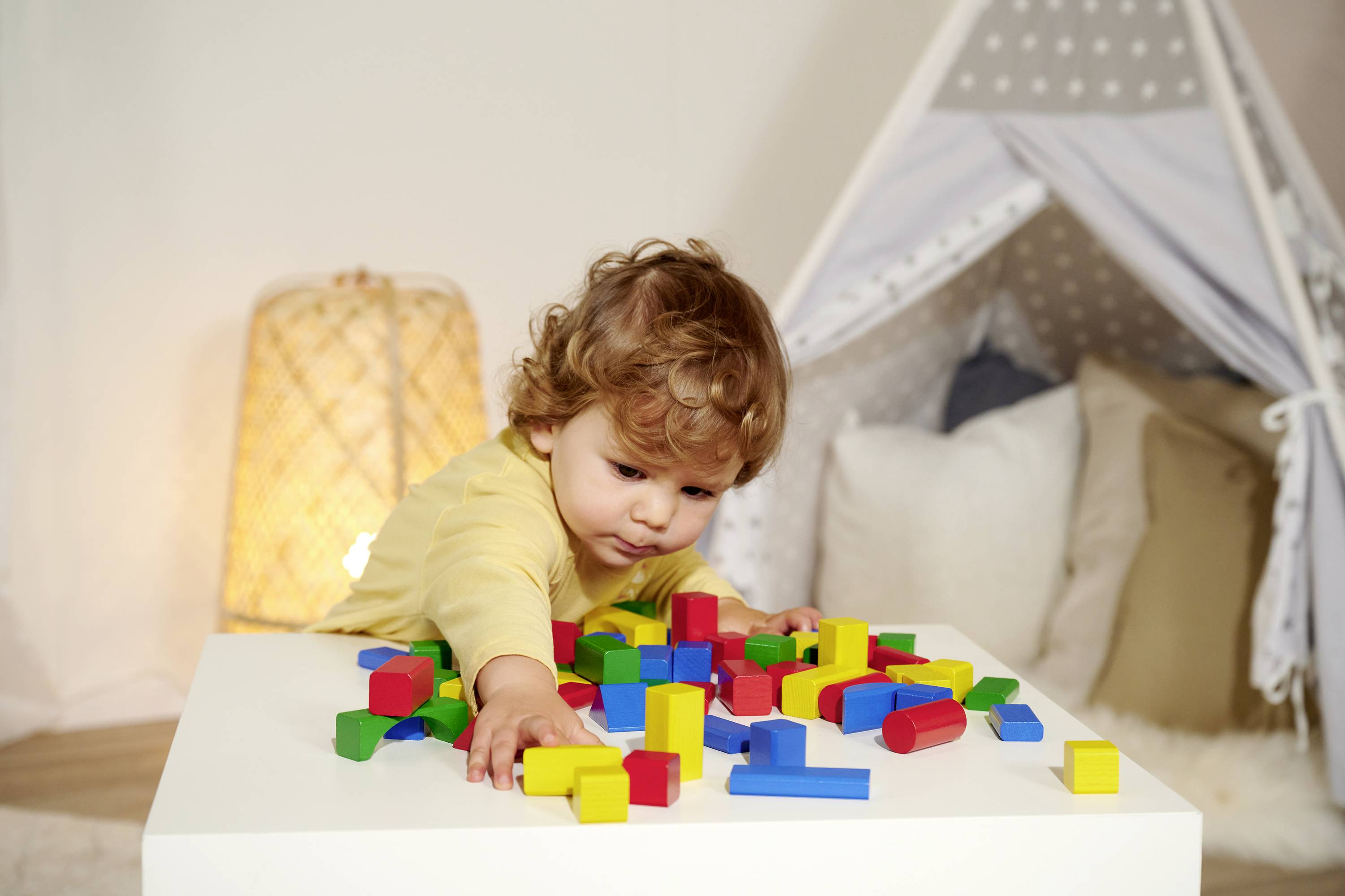 A child is playing with coloured building blocks on a table, with a tent and lamp in the background.