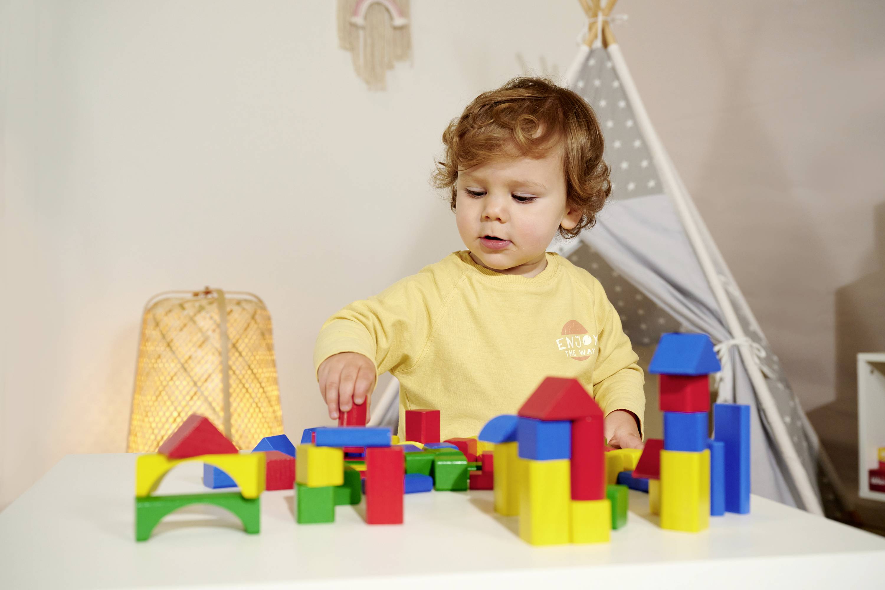 A toddler plays intently with colourful wooden building blocks on a table. In the background, a tent stands and a lamp glows warmly.