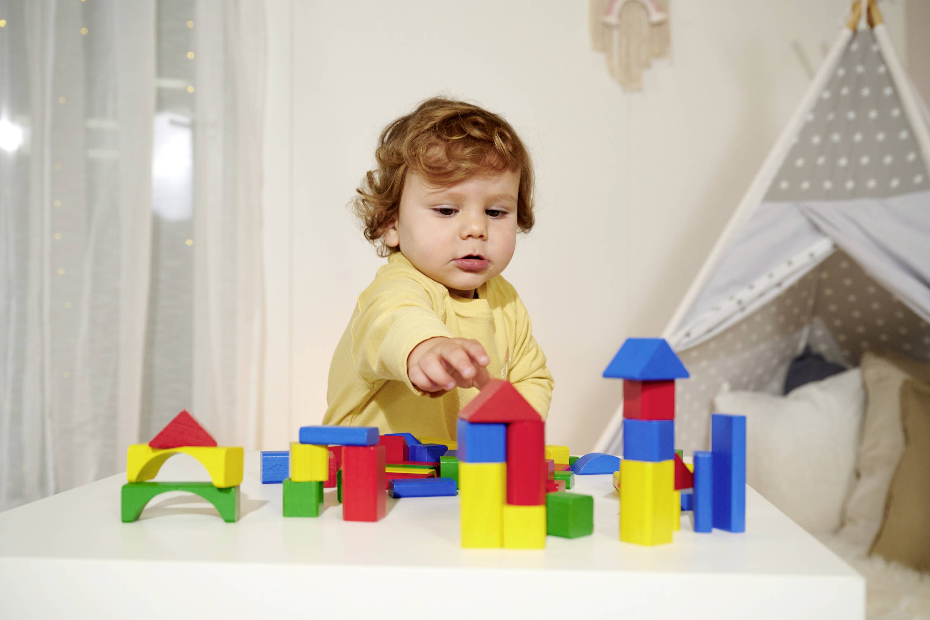 A toddler in a yellow top is playing with colourful wooden building blocks on a table. A play tent is visible in the background.
