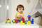 A toddler in a yellow top is playing with colourful wooden building blocks on a table. A play tent is visible in the background.