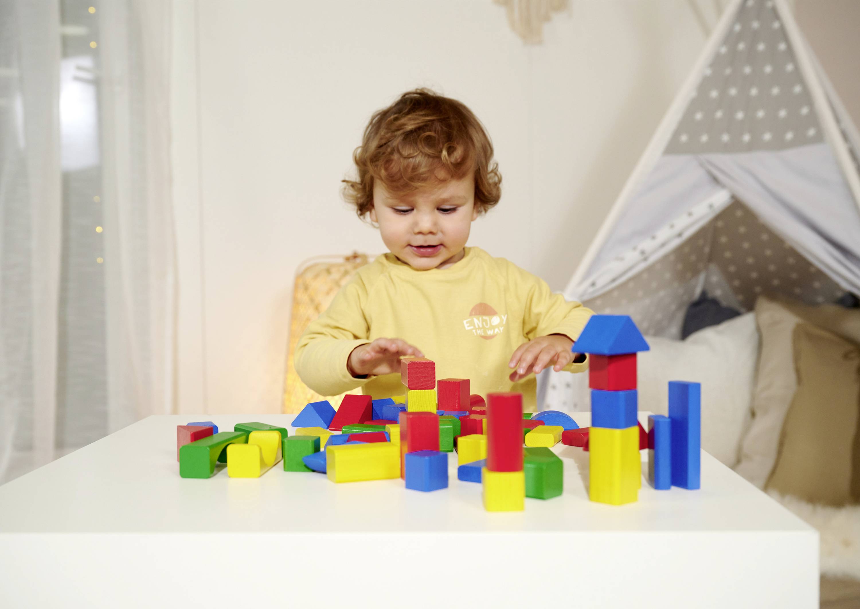 A toddler is playing with colourful building blocks on a table. In the background, a tent and sofa create a cosy play environment.