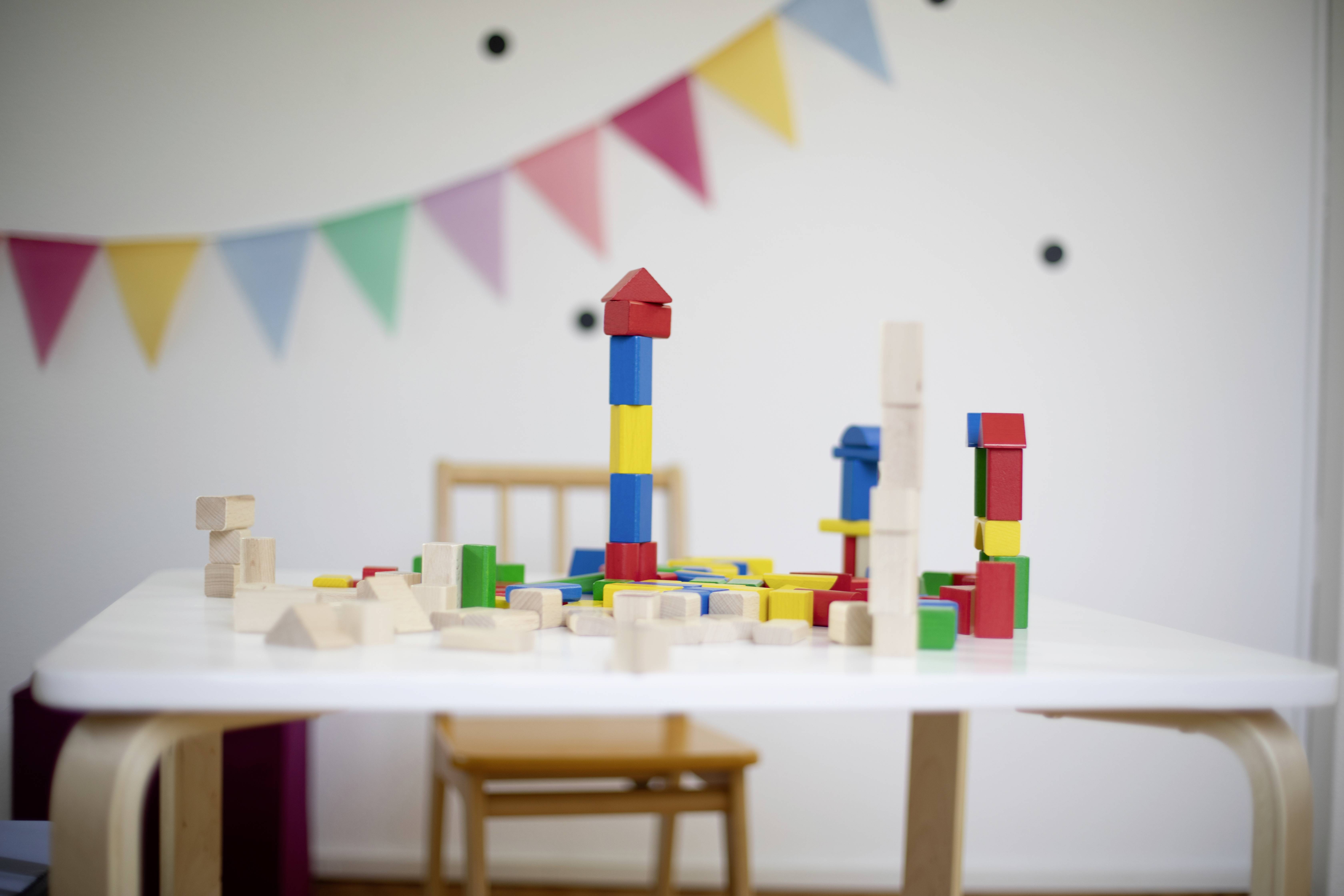 A table with colourful building blocks and wooden blocks on it, with a colourful bunting chain in the background. Playroom atmosphere.