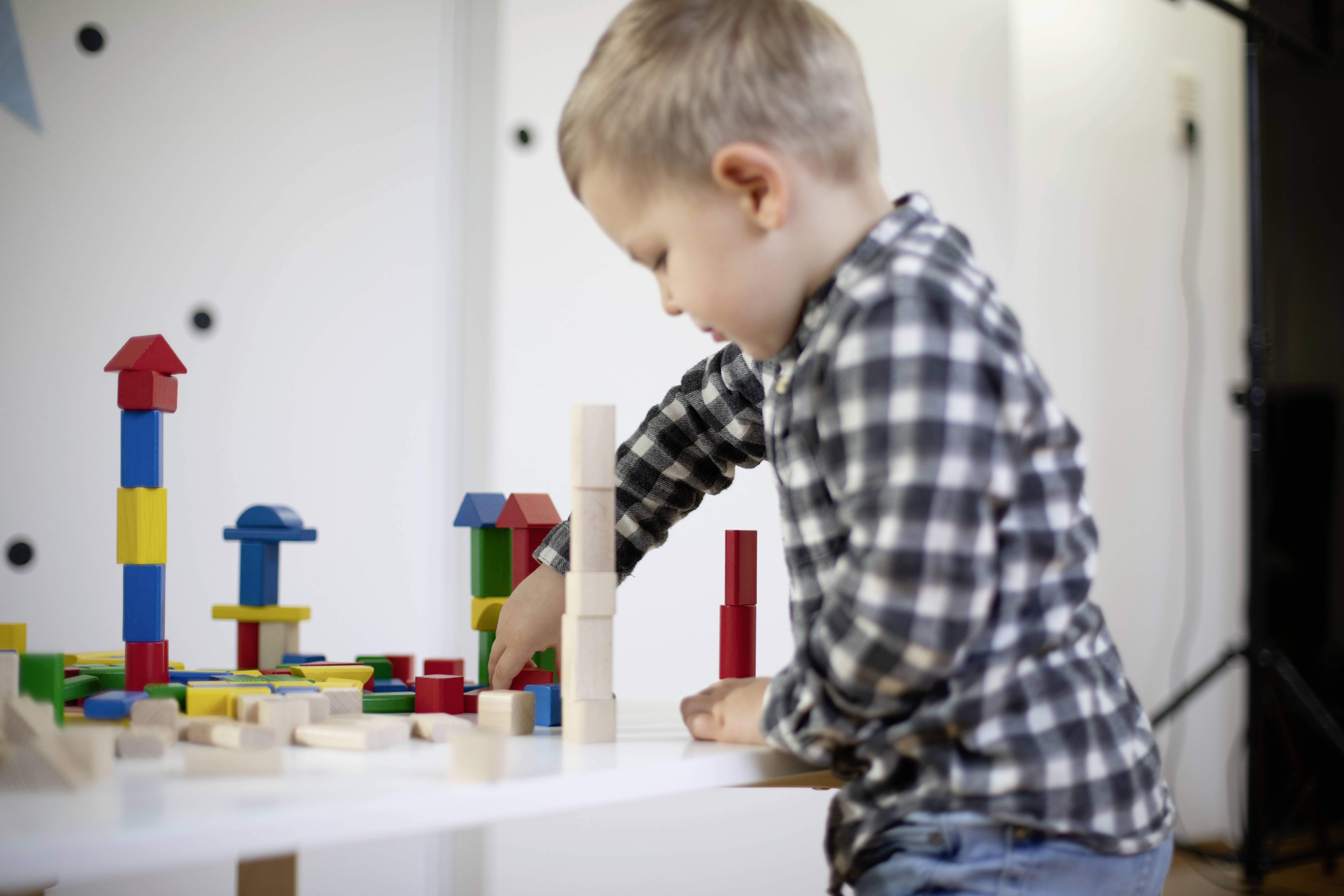 A child is playing with colourful building blocks on a table. The boy is wearing a checked shirt and is concentrating on constructing a structure.