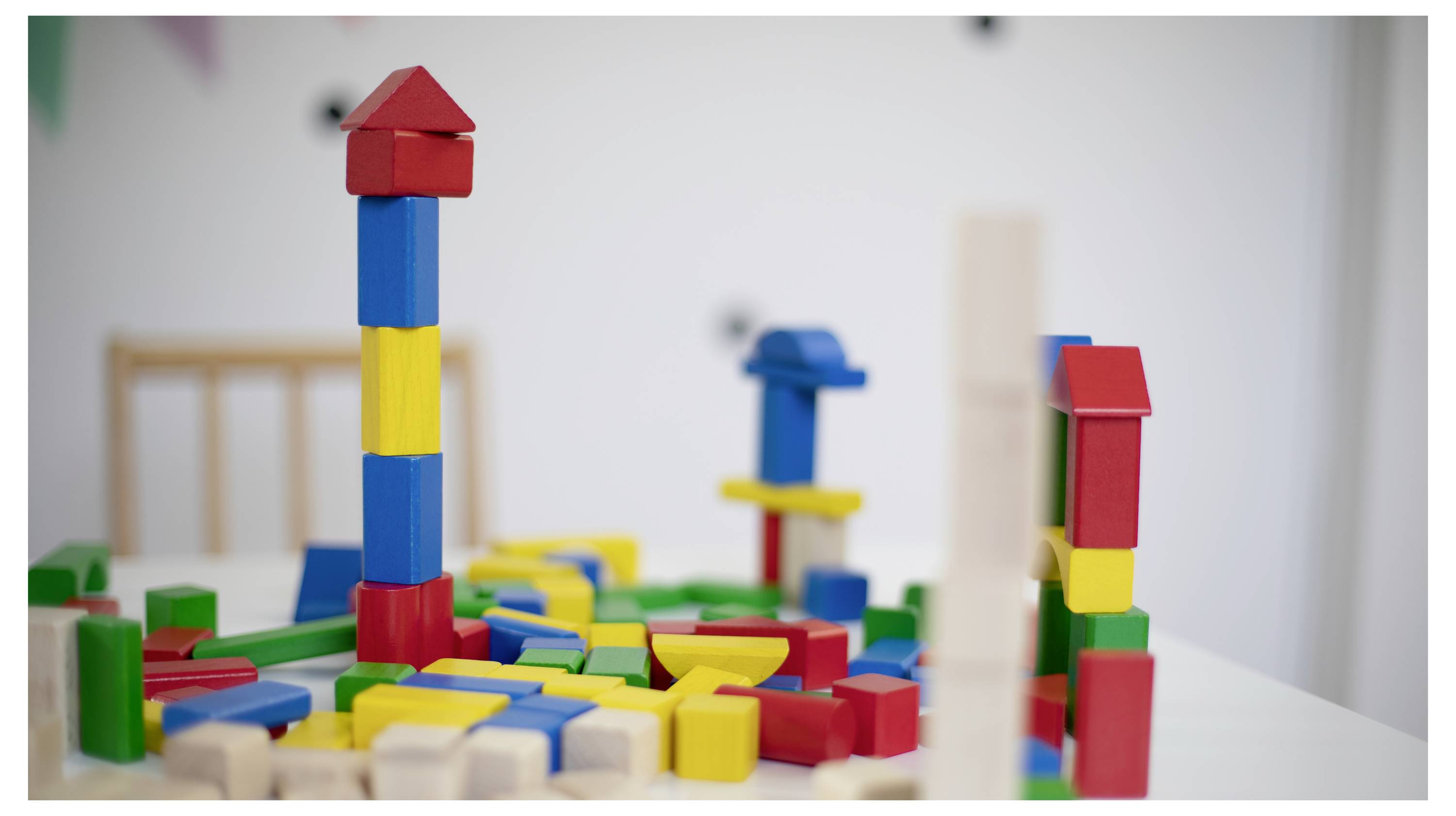 Colourful wooden building blocks are stacked on a table. A tall tower stands in the foreground. A chair is visible in the background.