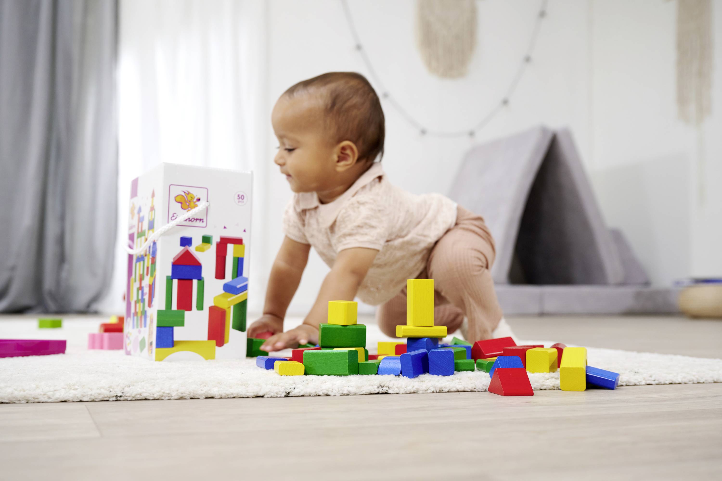 A toddler is playing on a carpet with colourful building blocks. A grey play tent structure is visible in the background.
