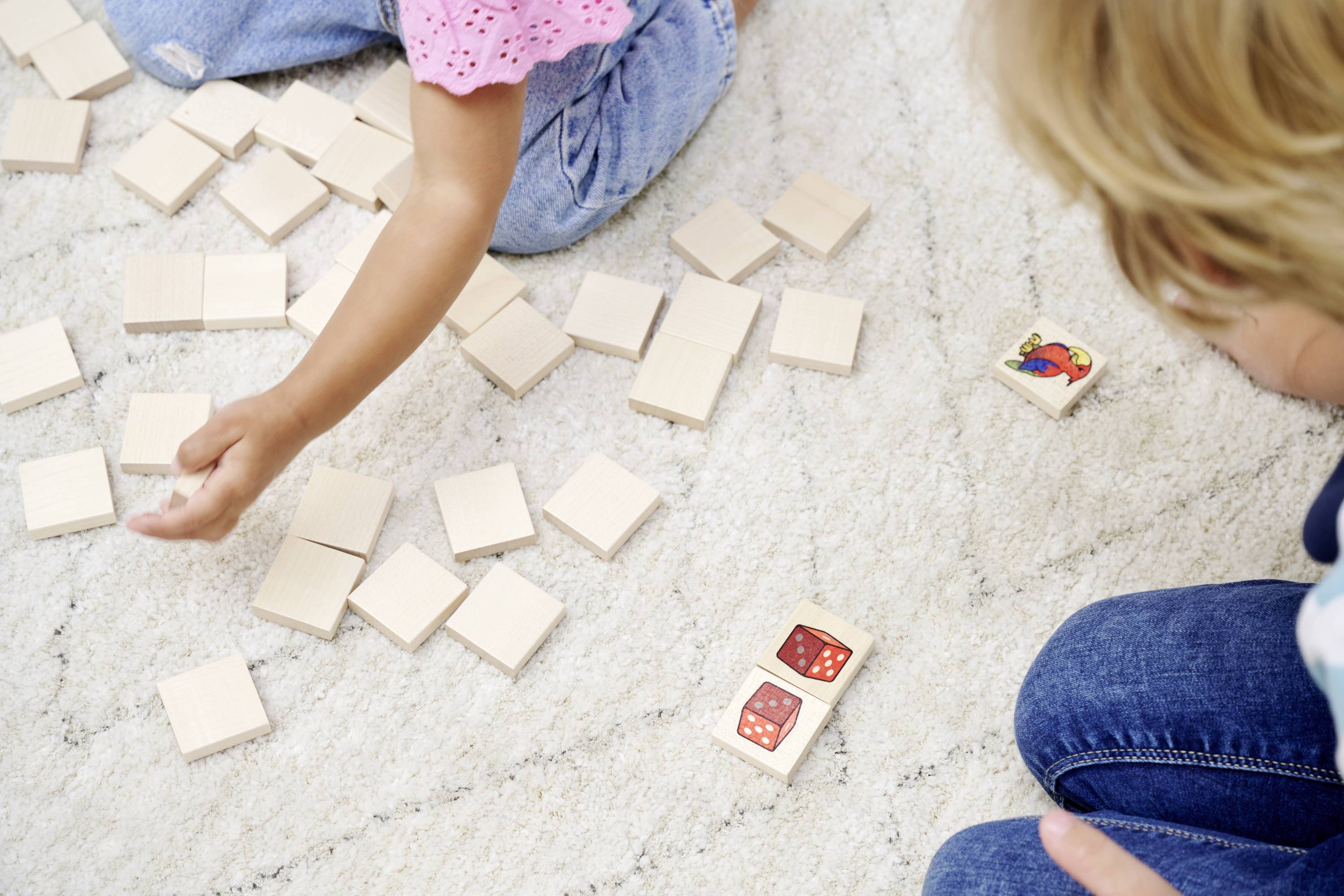 A child is playing on a rug with a wooden memory game. Another child is sitting nearby and watching.