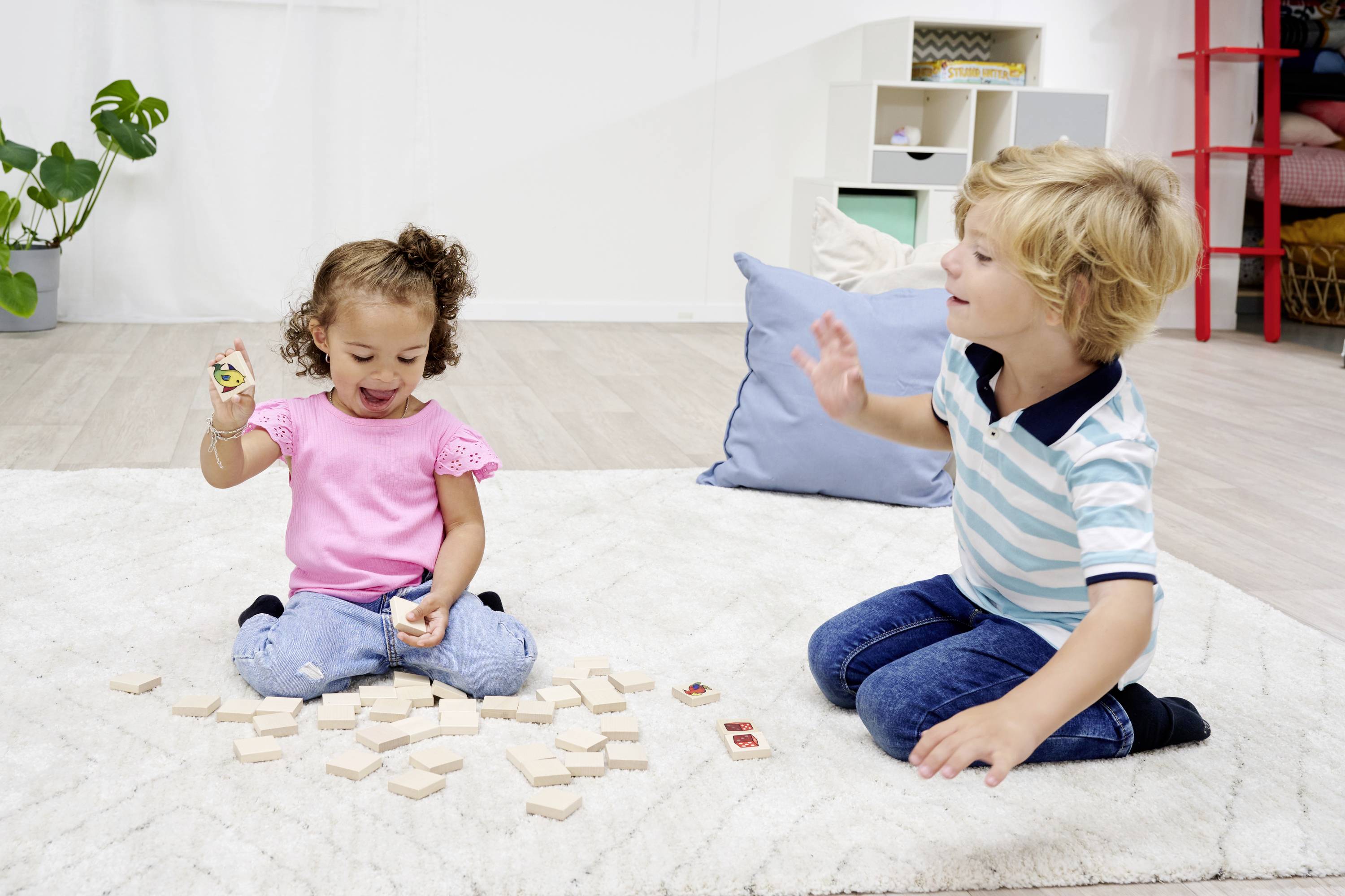 Two children are playing on a rug with building blocks. A girl is lifting a block, a boy is sitting beside her. Shelves and a plant are visible in the background.
