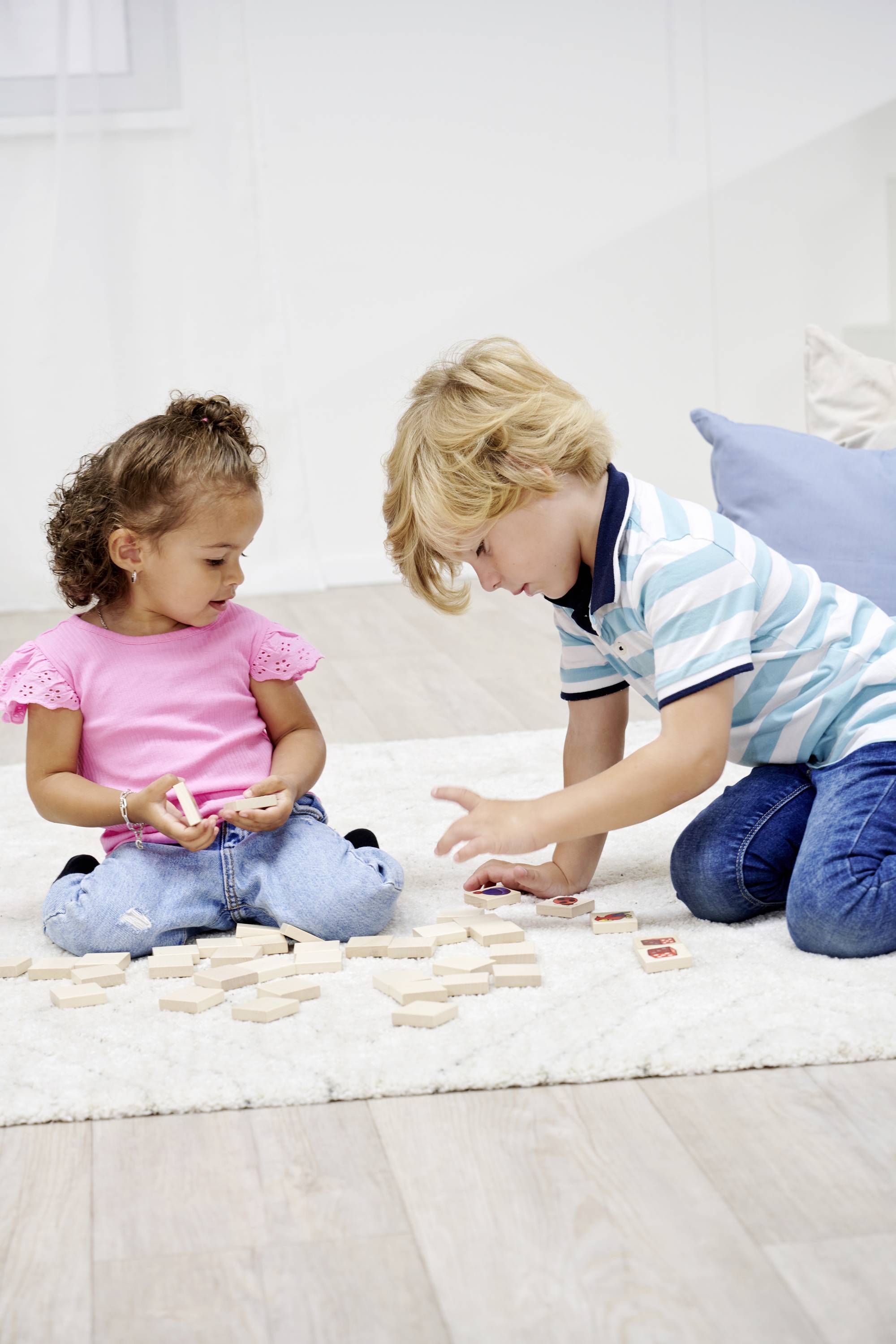 Two children are playing on a white carpet with wooden blocks. The girl is wearing a pink T-shirt, the boy a striped shirt.