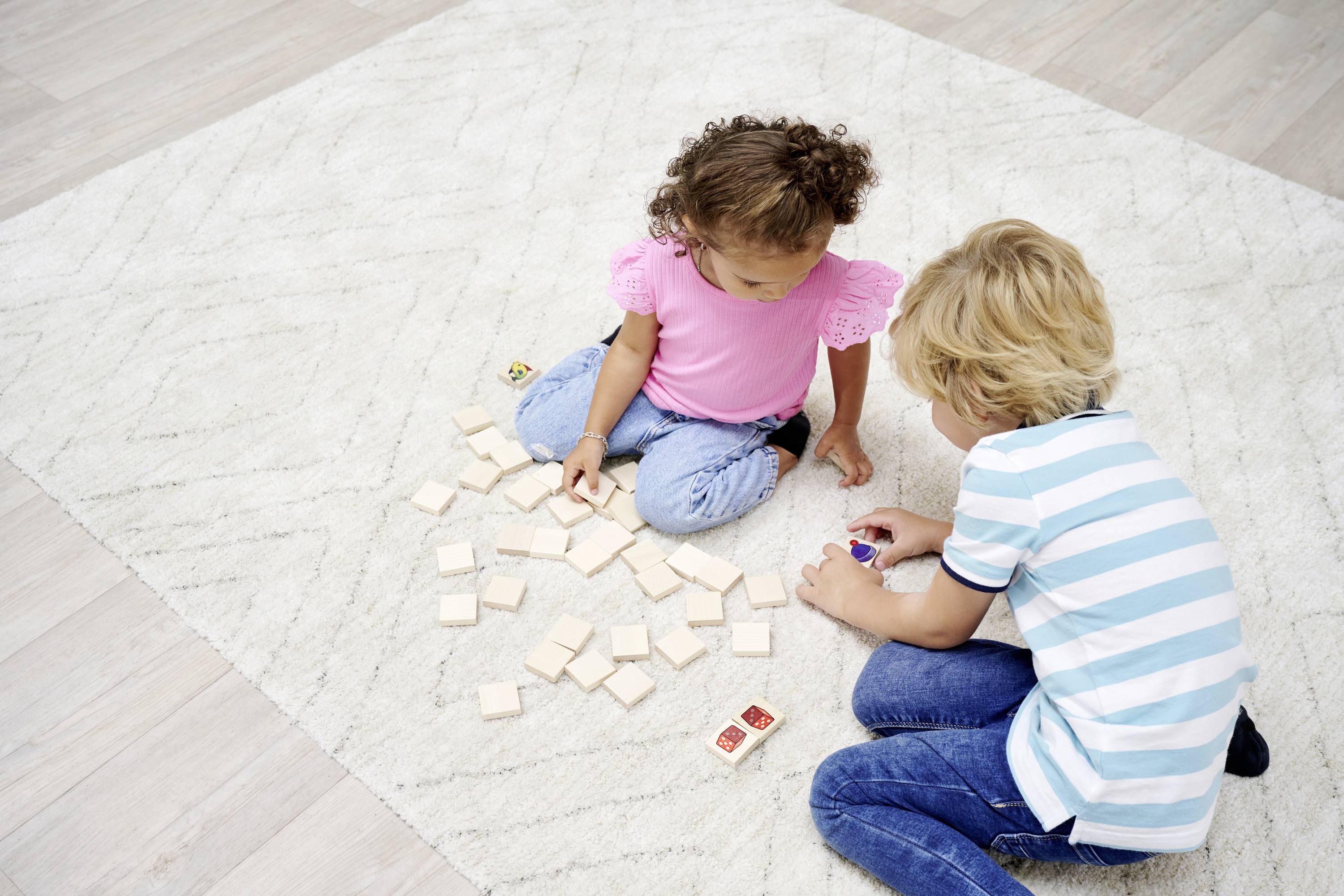 A girl and a boy are sitting on a rug and playing with square cards. They are focused on the game.