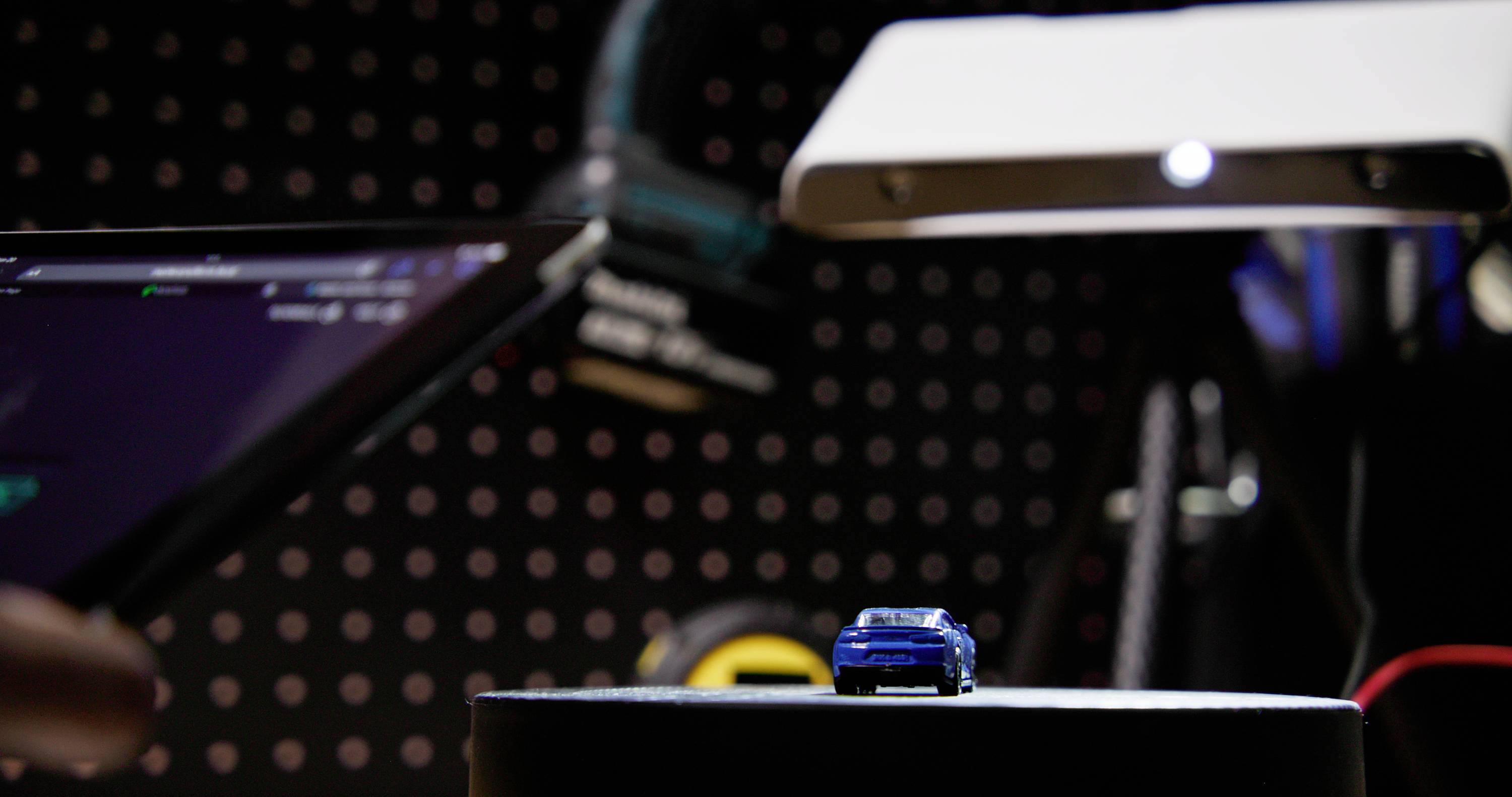 A miniature car is photographed under a lamp. A monitor and technical equipment are visible in the background, suggesting a studio environment.