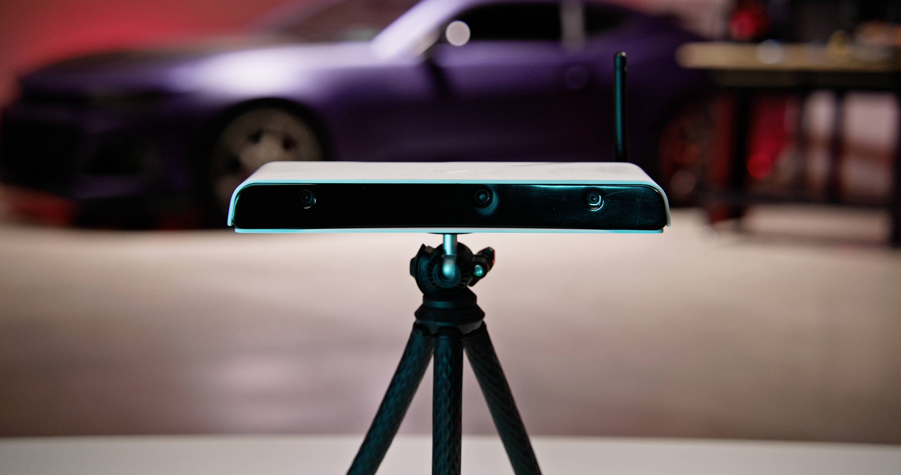 A white device with an antenna mounted on a tripod stands in the foreground, with a blurry car in a workshop behind it.