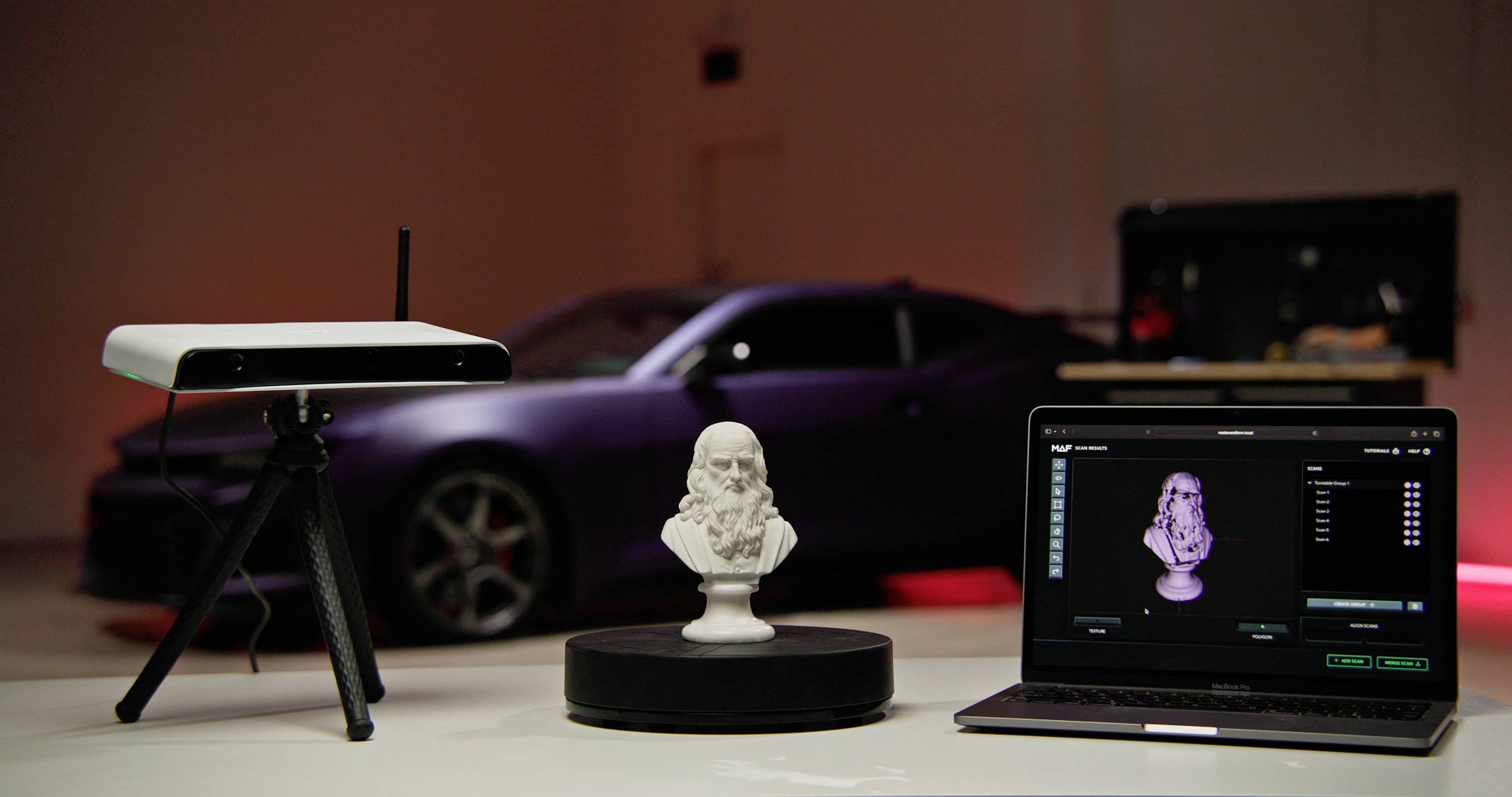 A 3D scanner is scanning a bust on a turntable, with the results displayed on a laptop. A sports car is positioned in the background.