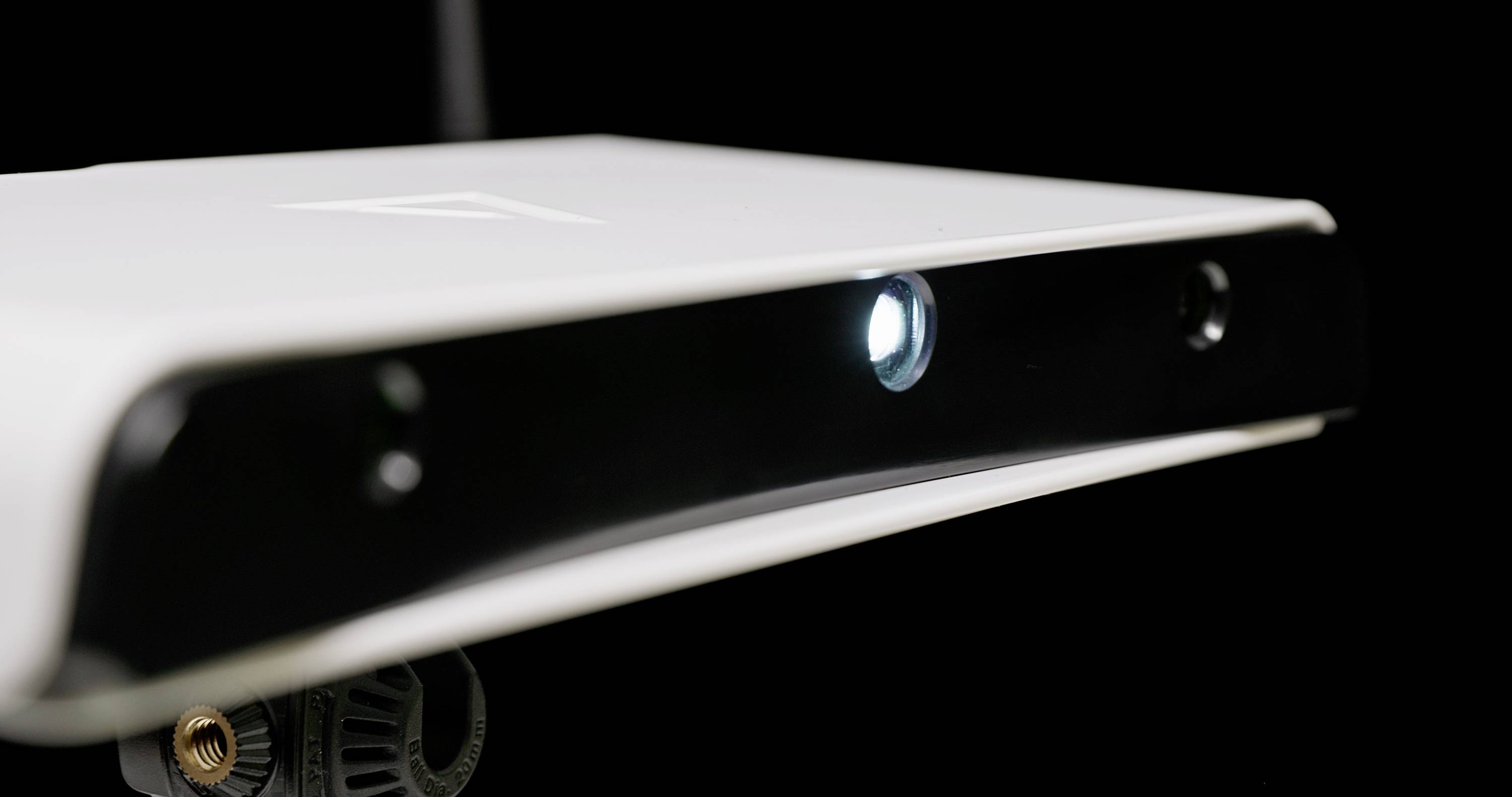 A white electronic device with a black front panel and a glowing sensor in the centre, possibly a camera or scanner, against a black background.