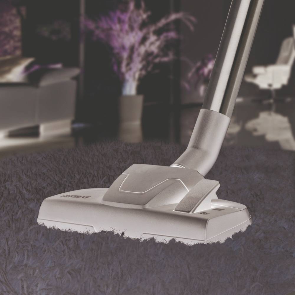 A hoover is vacuuming a fluffy carpet. Blurry furniture and plants can be seen in the background.