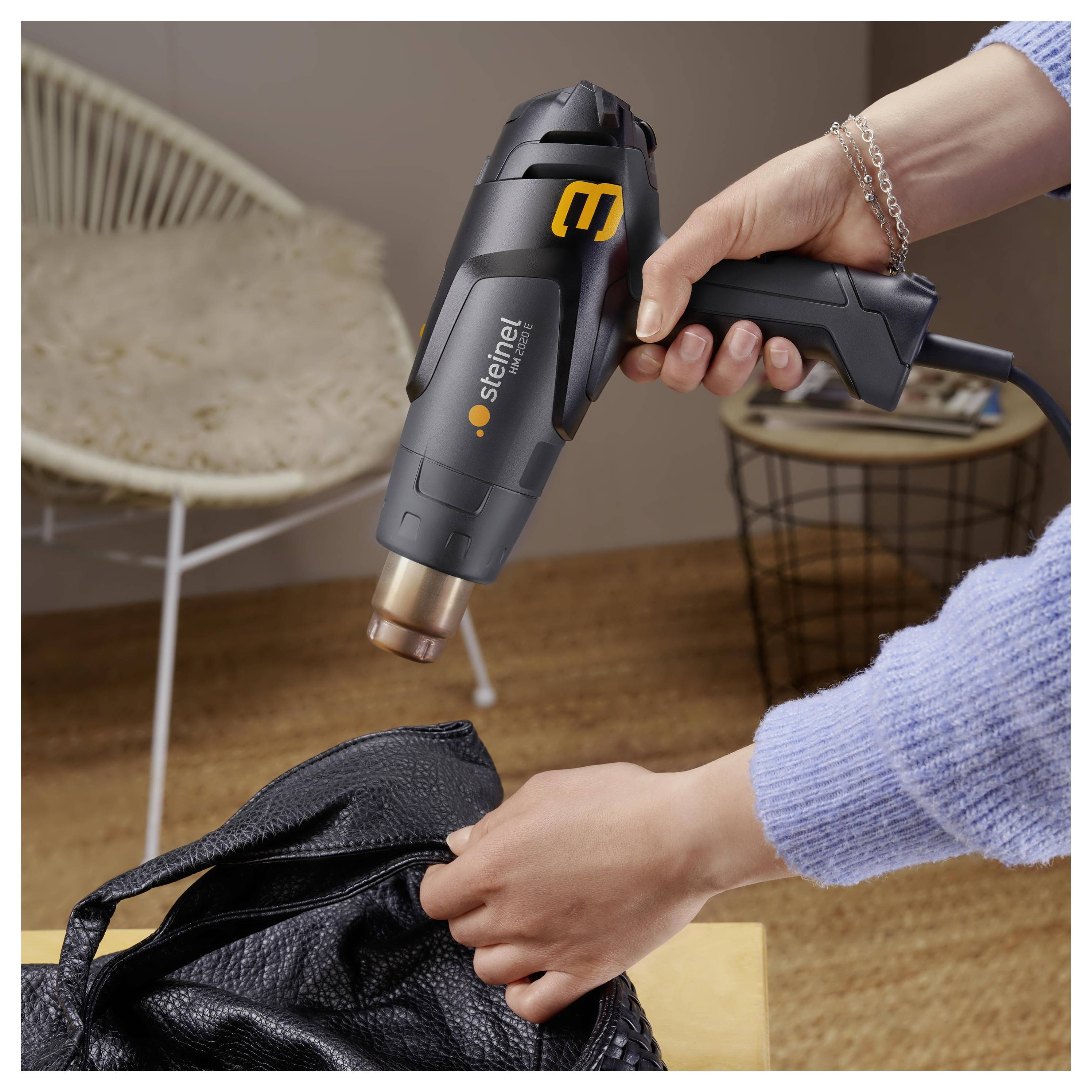 Handheld Steinel hot air gun being used to heat a black bag or material in a home setting.