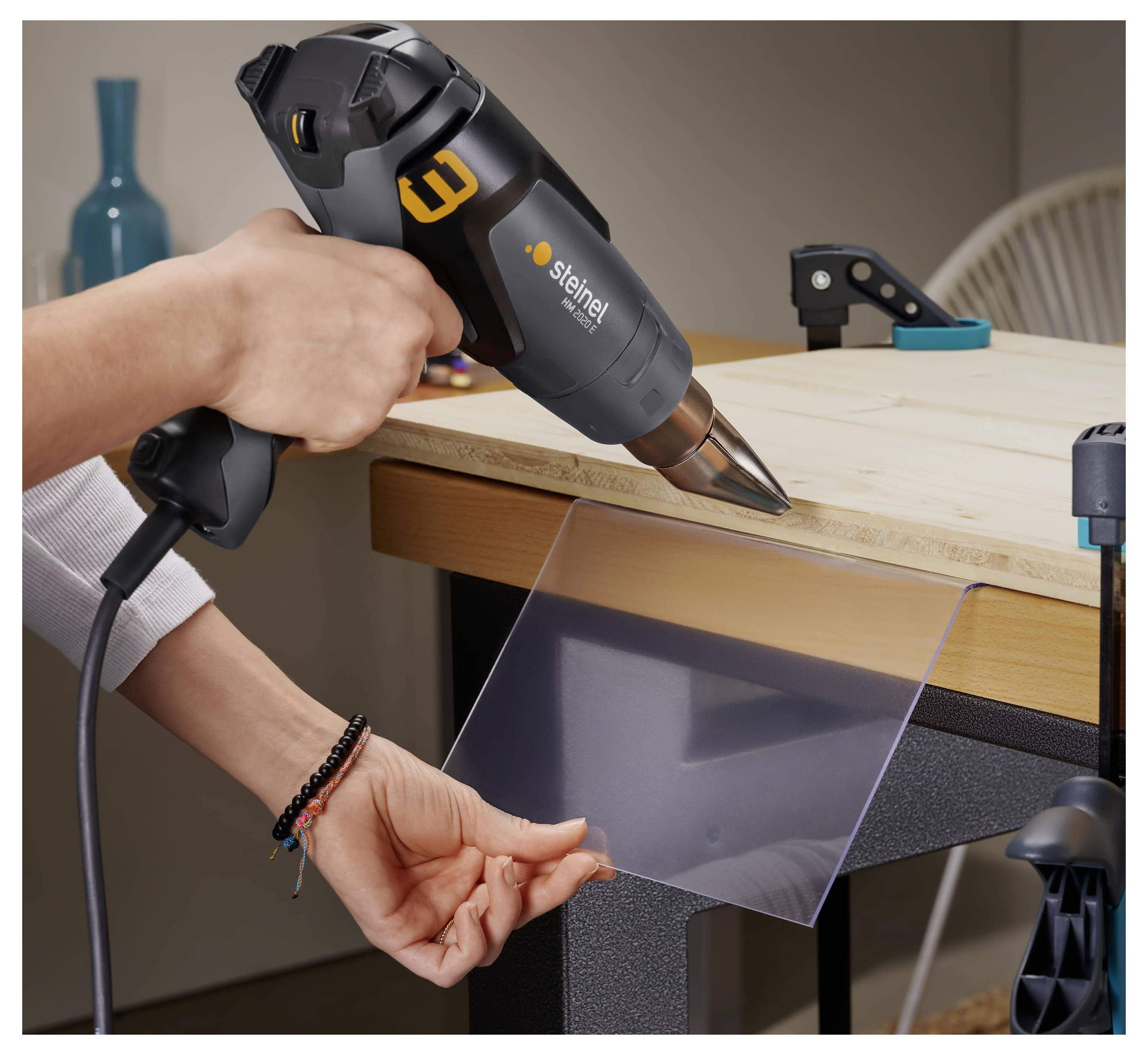 Person uses a heat gun to bend a clear plastic sheet over a workbench.