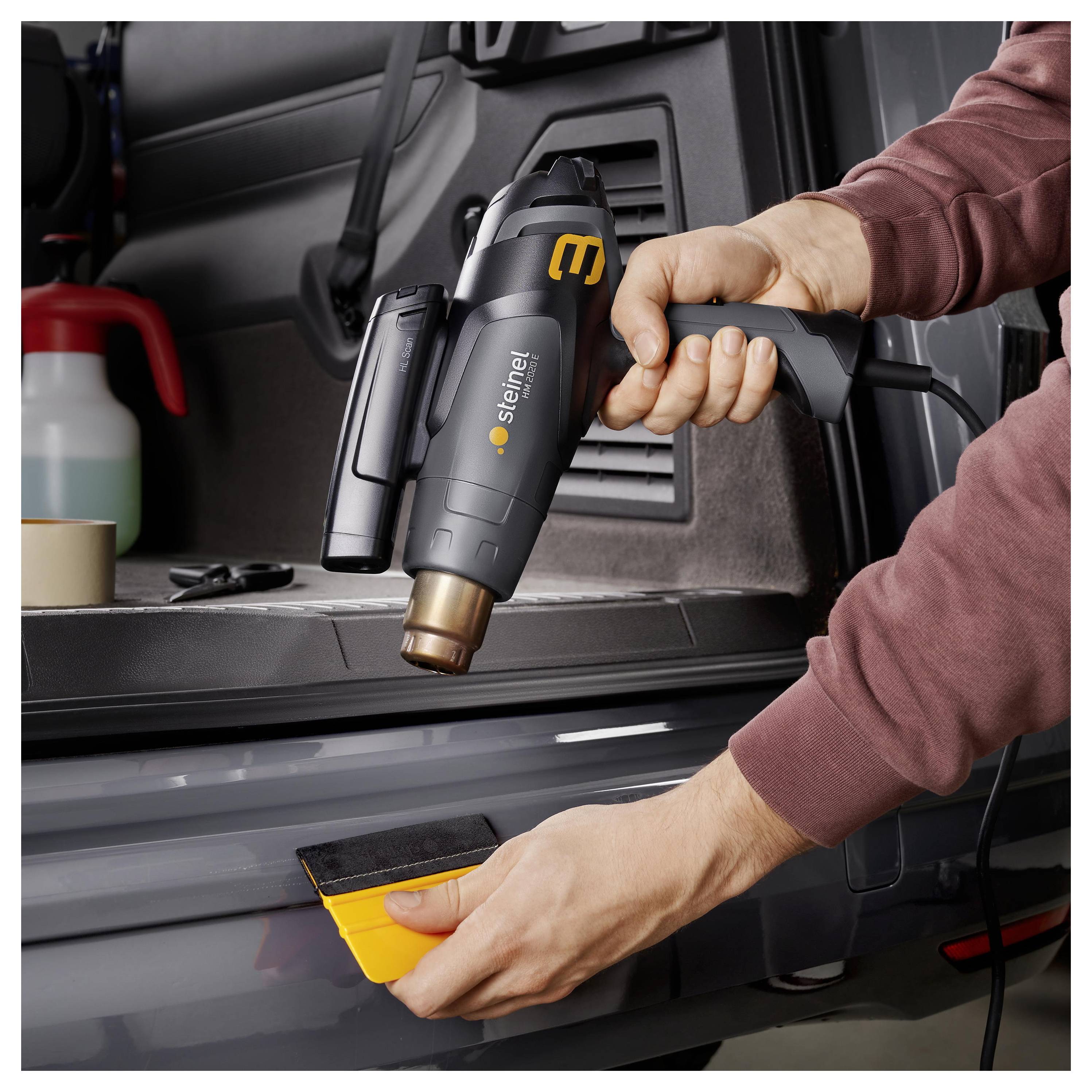 Person using a heat gun to apply a yellow vinyl film to a car surface inside an open trunk.