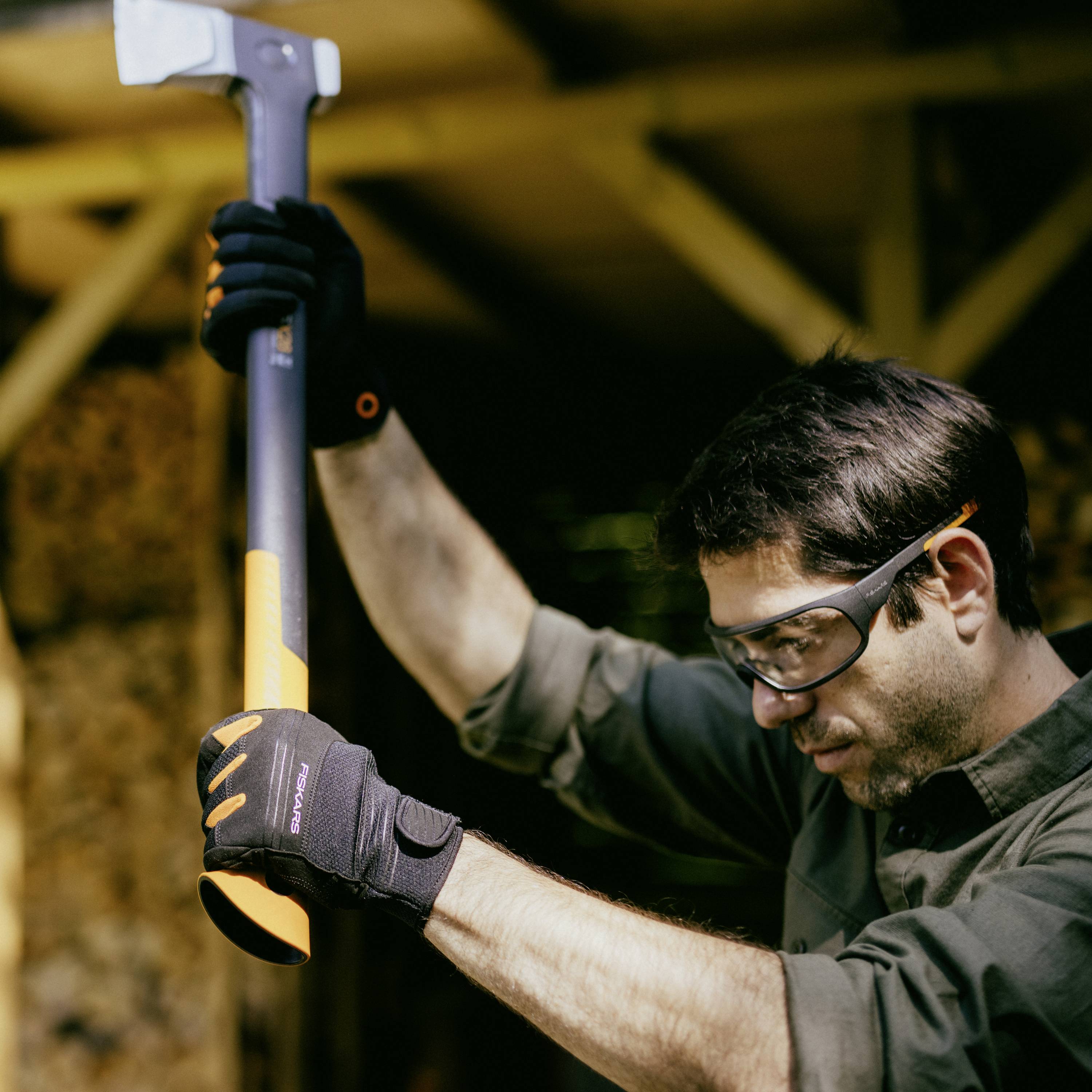 A man wearing safety glasses and gloves is swinging an axe, ready to strike a wooden block. Stacks of wood can be seen in the background.