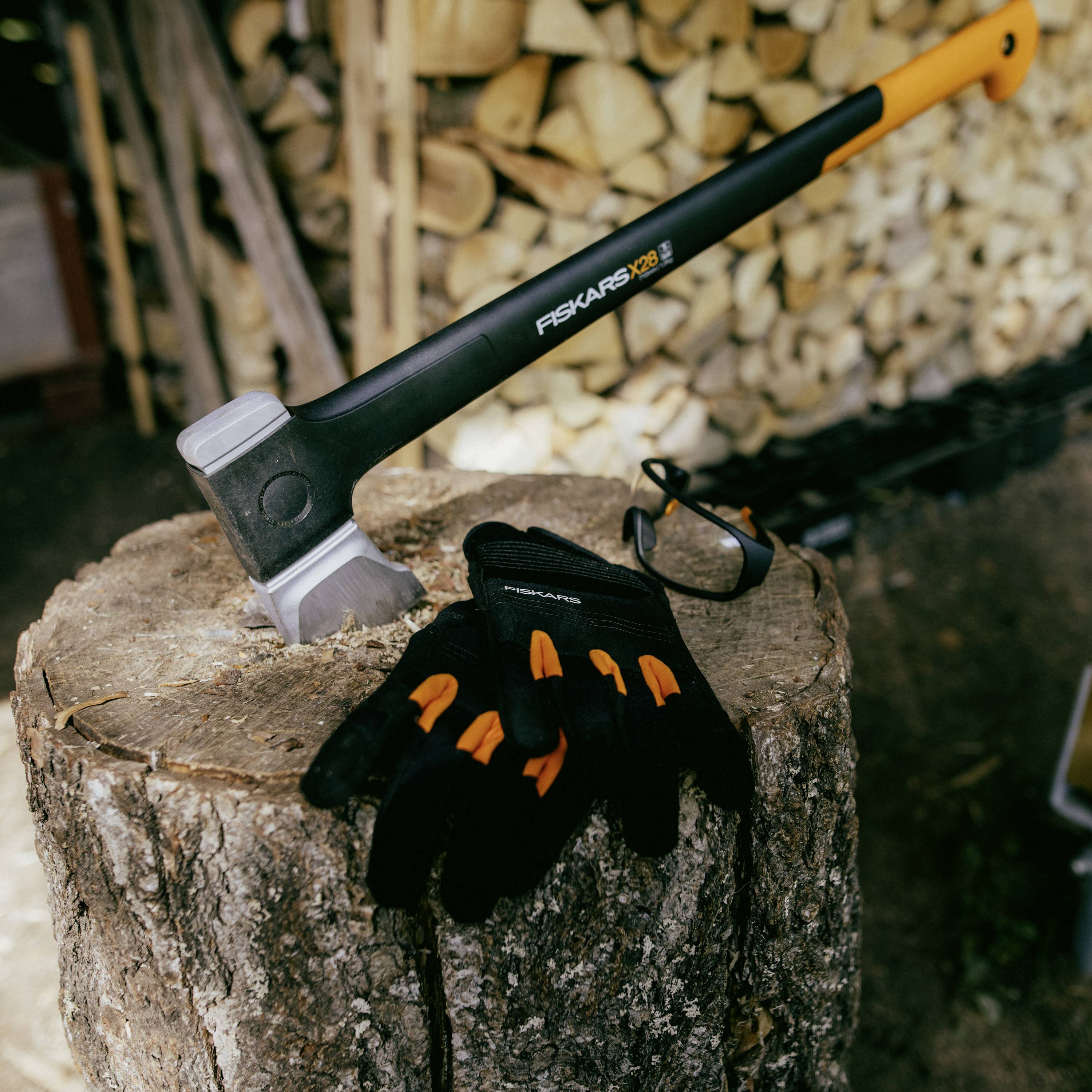 An axe with an orange handle rests on a tree stump, next to black gloves with orange details, with stacked wood in the background.
