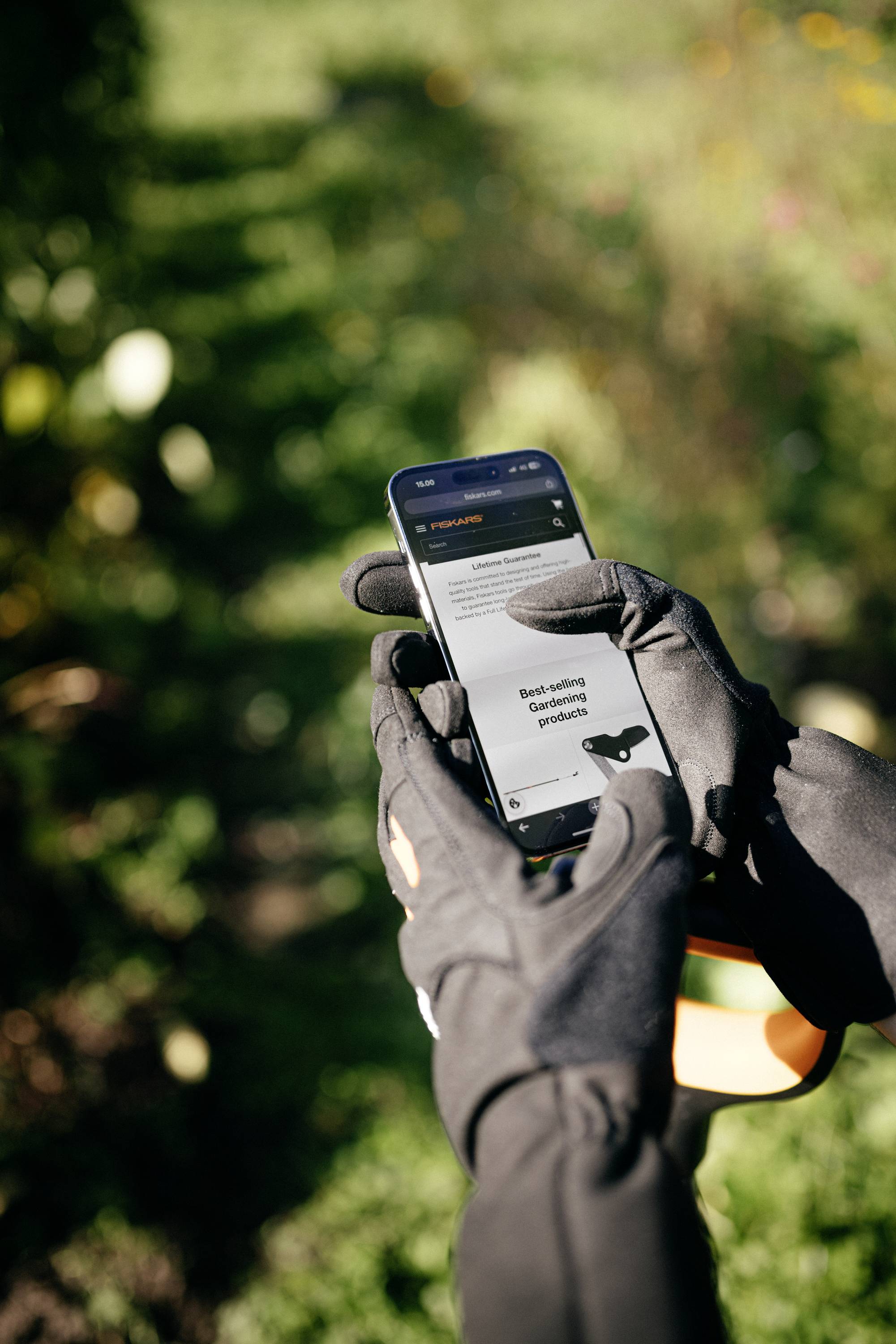 A person wearing black gloves holds a smartphone outdoors, displaying an open webpage. The background is green and blurred.