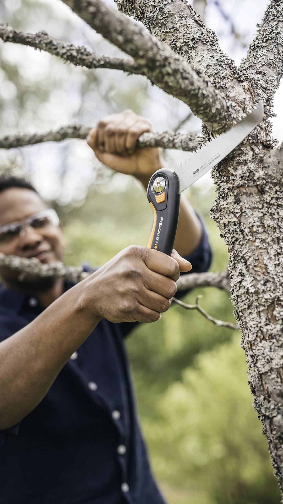 A person is sawing a branch off with a pruning saw. The background shows a wooded, green area.