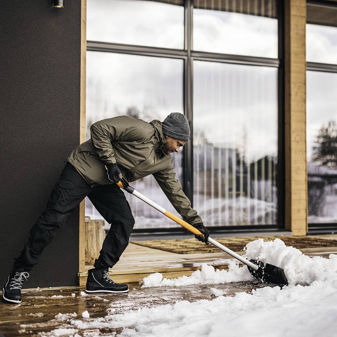 A man in winter clothing is shovelling snow on a wooden terrace in front of a modern house. Background: snowy landscape.