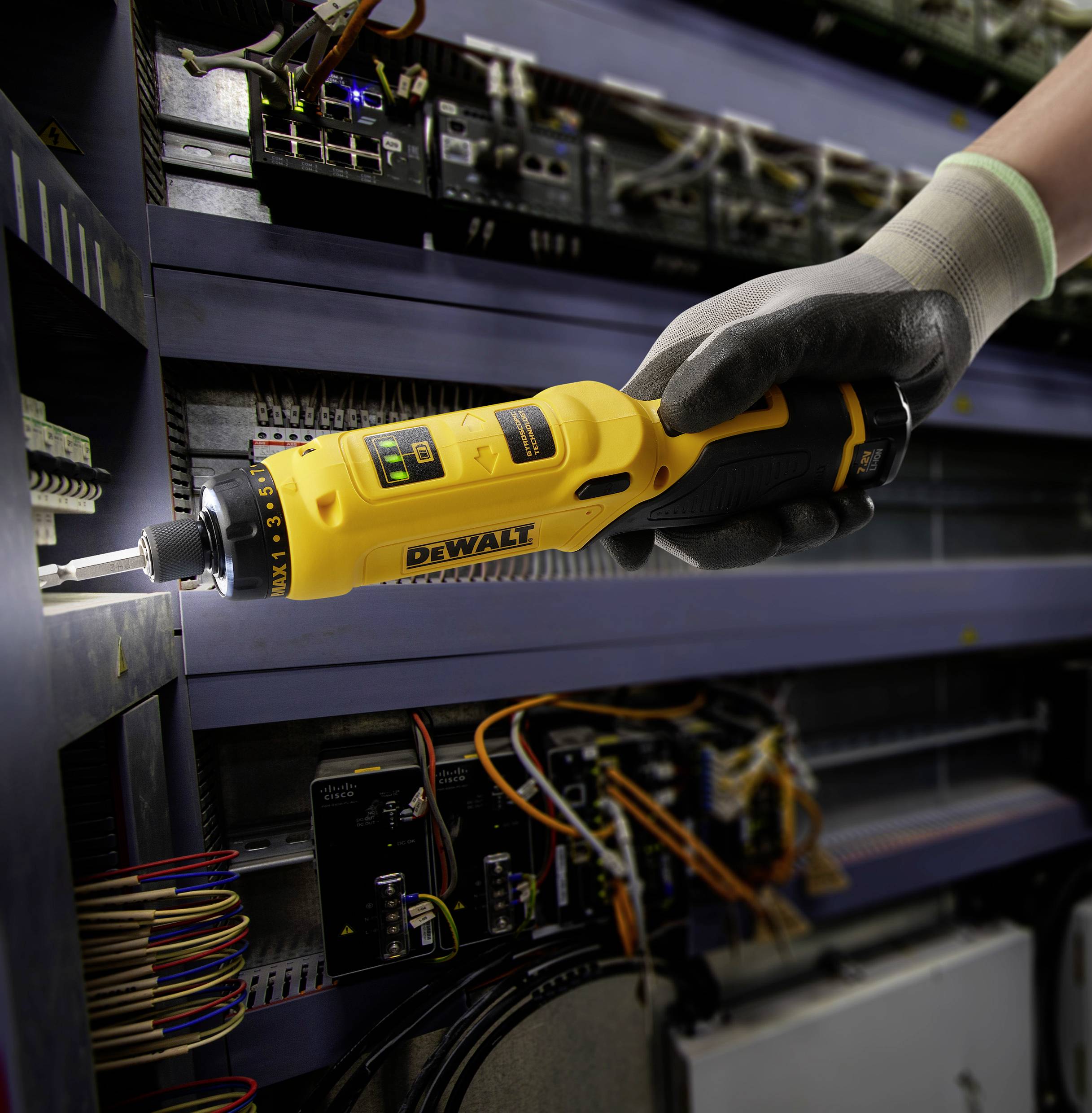 A person wearing work gloves is using a yellow cordless drill to secure or loosen cables on an electrical cabinet.