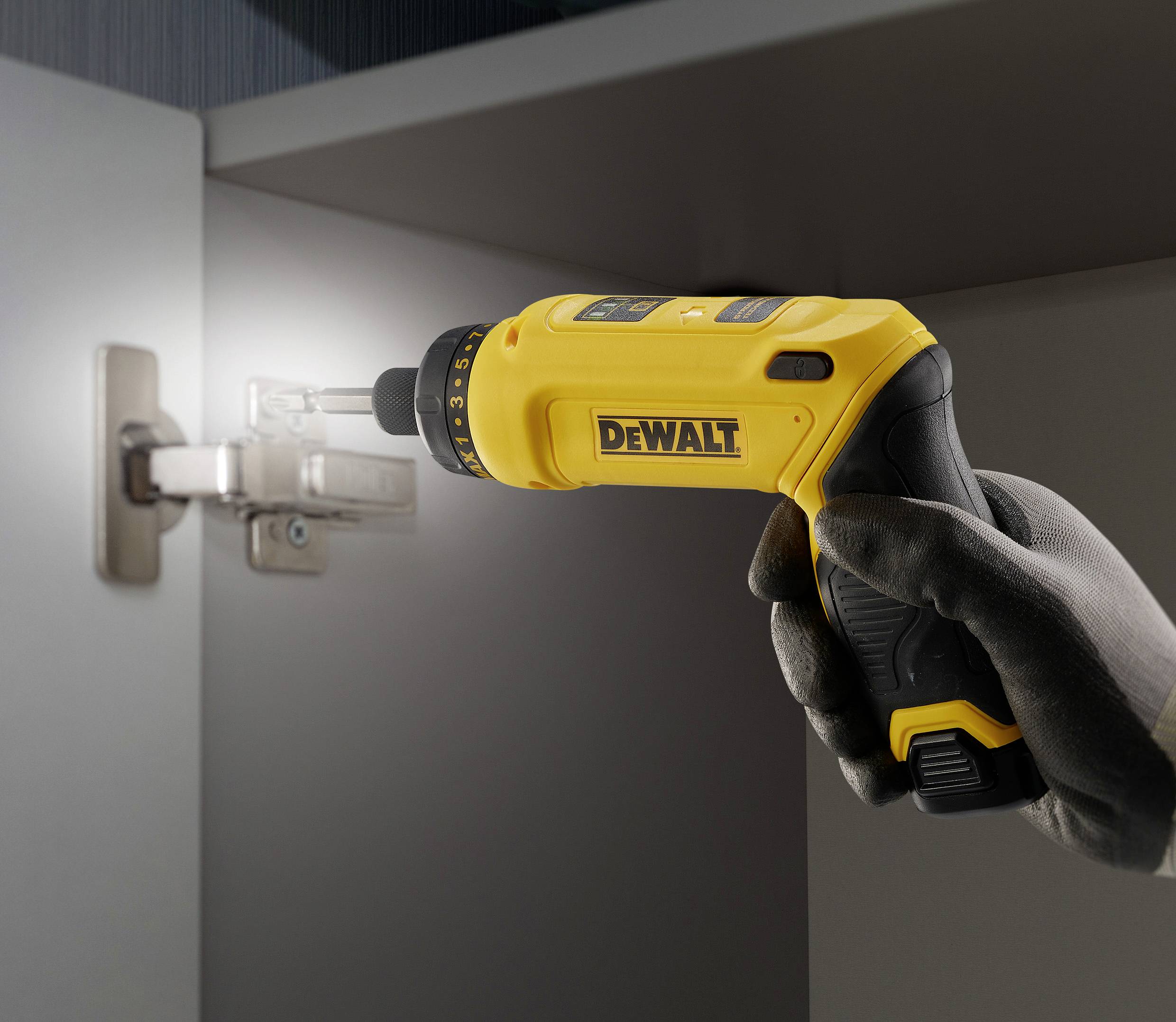 A gloved hand holds a yellow cordless drill, securing a hinge to a cabinet.