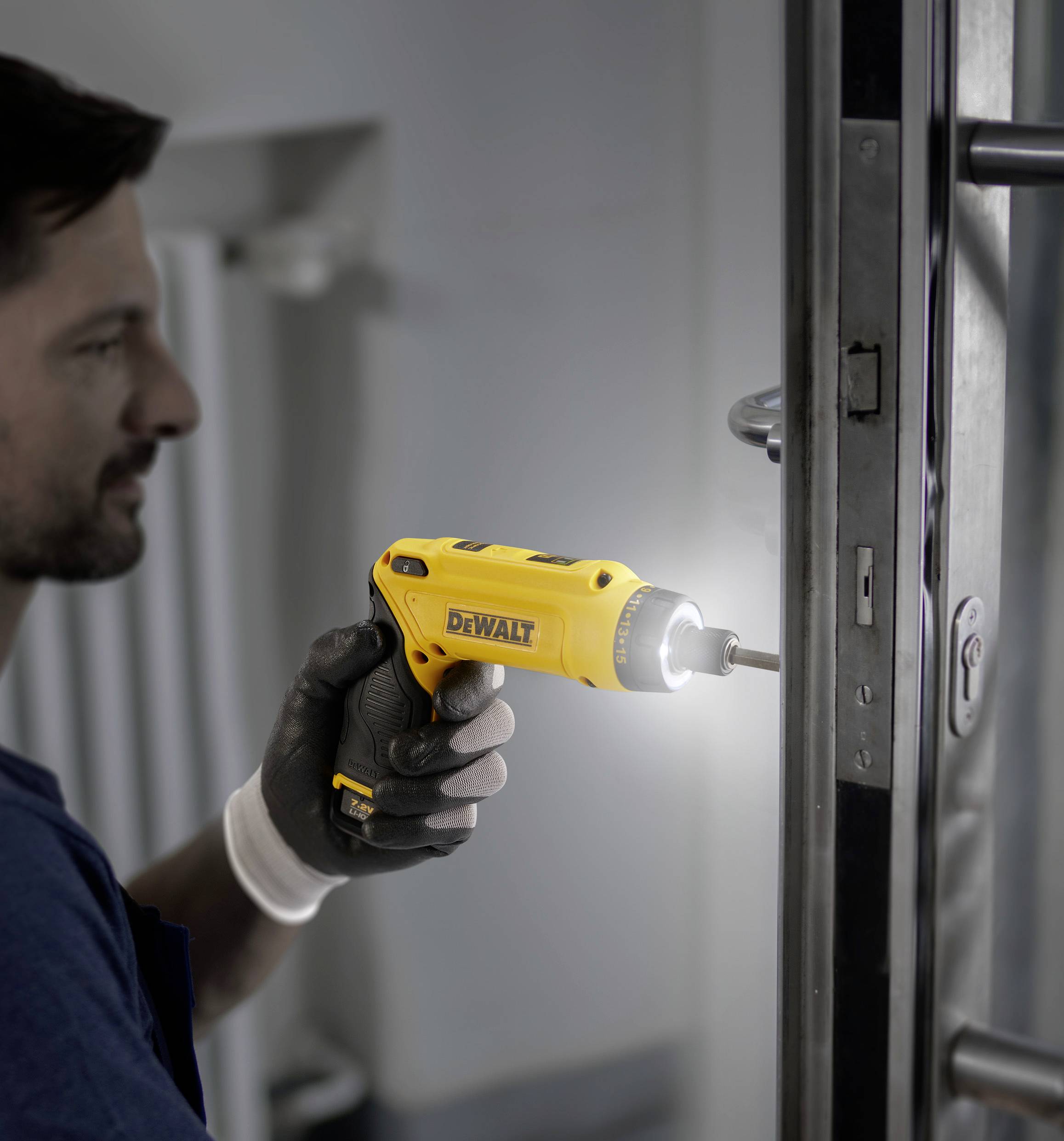 A man is using a yellow compact cordless screwdriver to repair a door. He is wearing work gloves and is focused on the task.