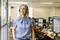 A woman wearing headphones smiles and looks out of an office window. Workstations are blurred in the background.