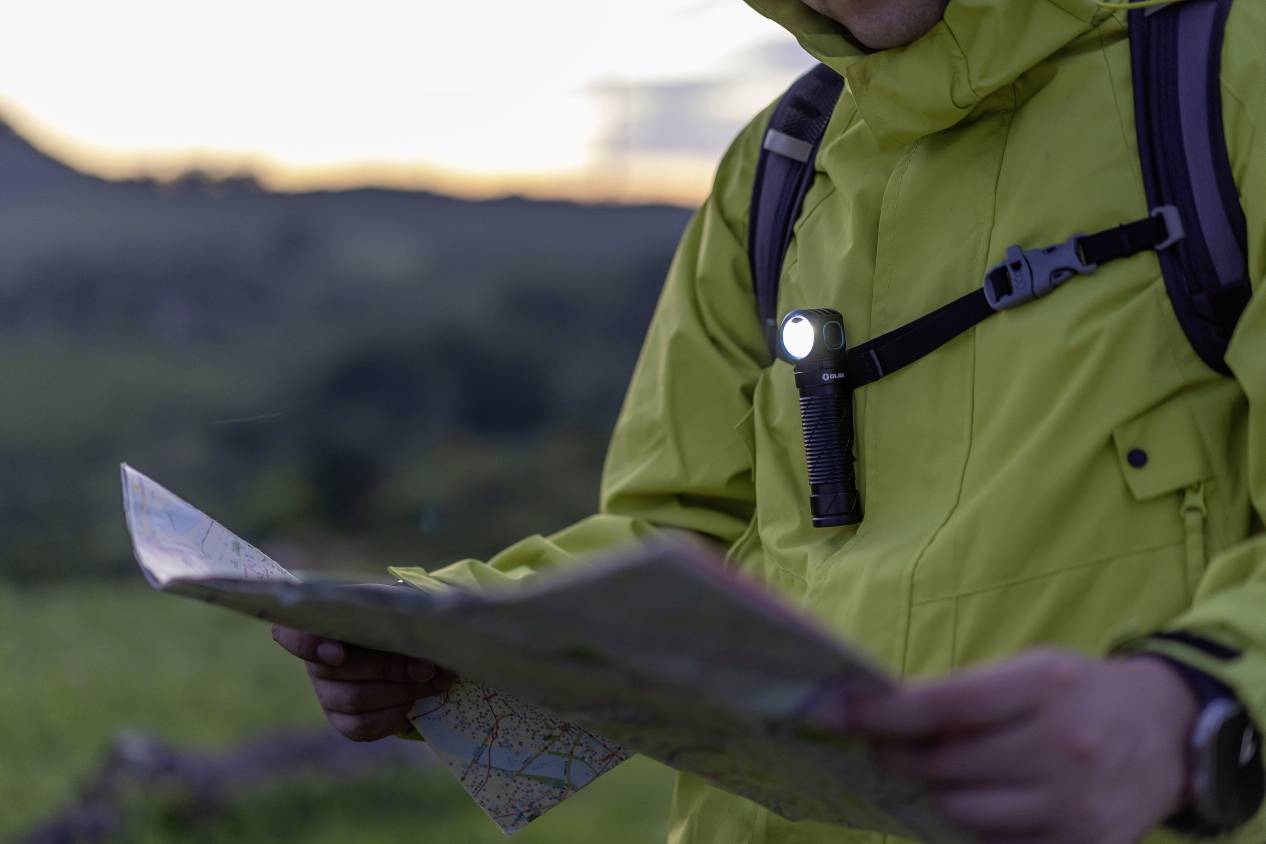 A person in a raincoat is reading a map while standing outdoors. It is twilight. A small torch is illuminating their chest.