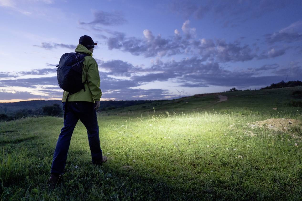 A person with a rucksack and head torch is hiking across a meadow towards the horizon at dusk, with a slightly cloudy sky in the background.