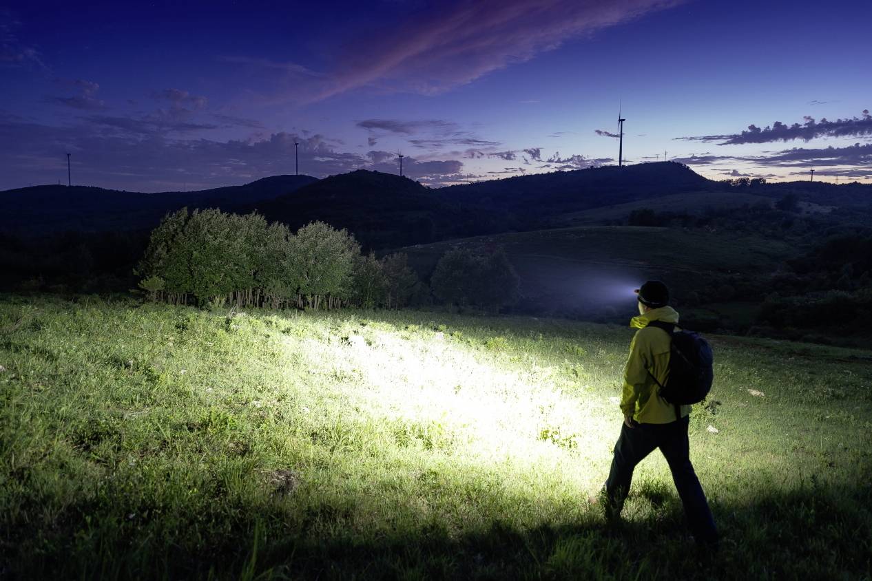 A person with a rucksack and head torch is walking across a field at night. Wind turbines and hills can be seen in the background.