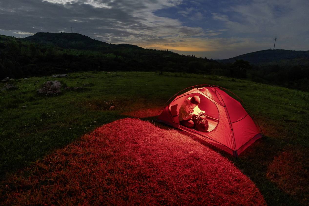A person sits in a red-lit tent on a meadow at sunset. Hills and clouds are visible in the background.