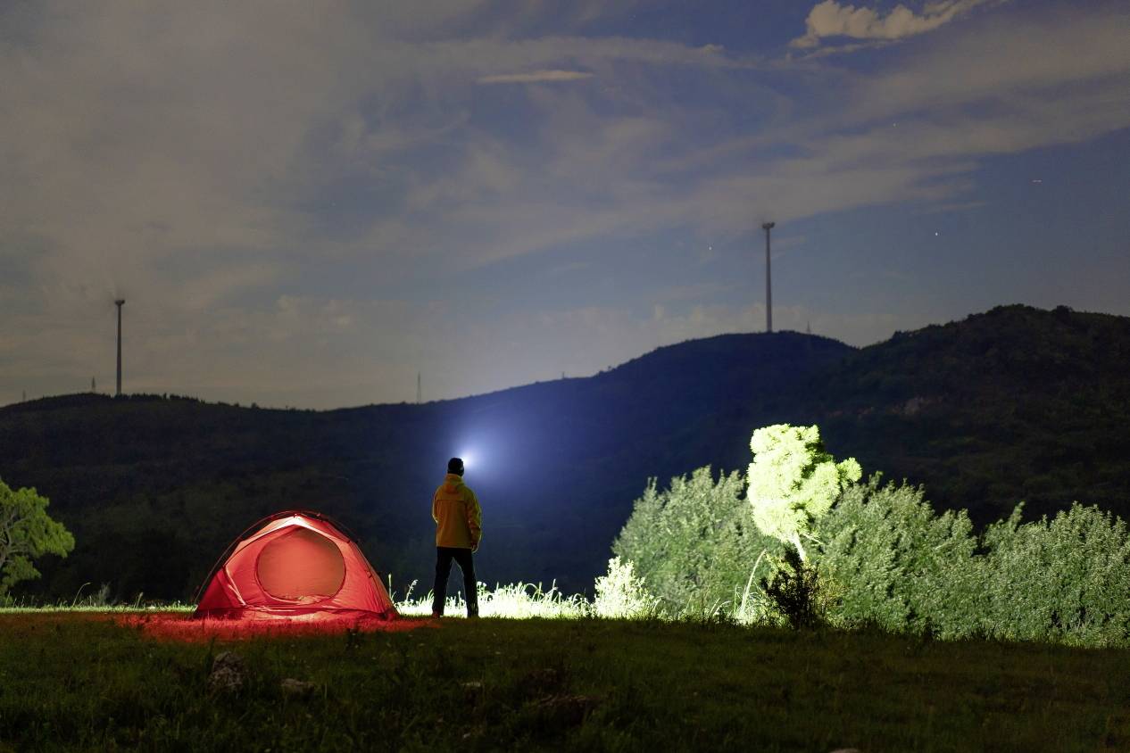 A person with a torch stands beside an illuminated tent on a meadow under a starry sky; hills and trees in the background.