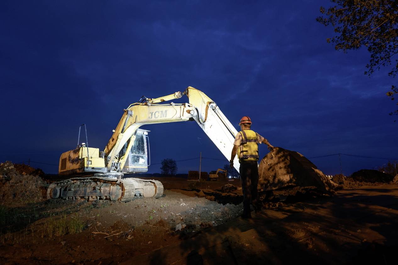 A construction worker stands beside an illuminated yellow excavator on a building site at night under a clear, dark sky.