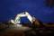 A construction worker stands beside an illuminated yellow excavator on a building site at night under a clear, dark sky.