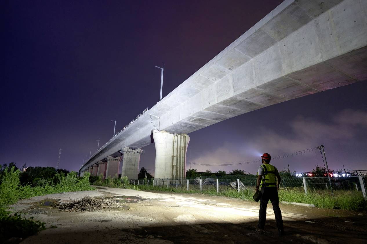 A construction worker wearing a hard hat stands at night beneath a partially completed, illuminated concrete bridge, surrounded by vegetation and lighting.
