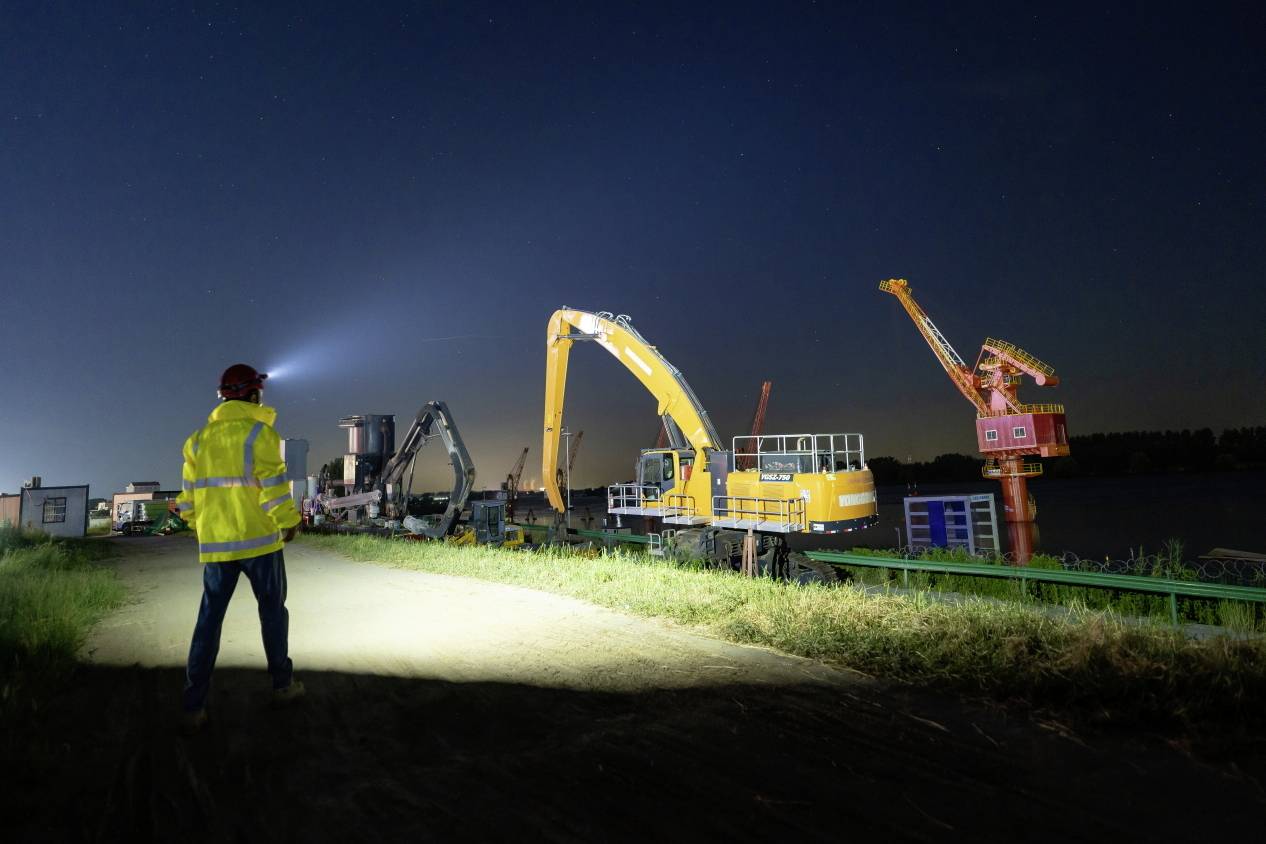 'A worker in a yellow protective suit stands by an illuminated river at night, while large construction machines operate on a building site.'