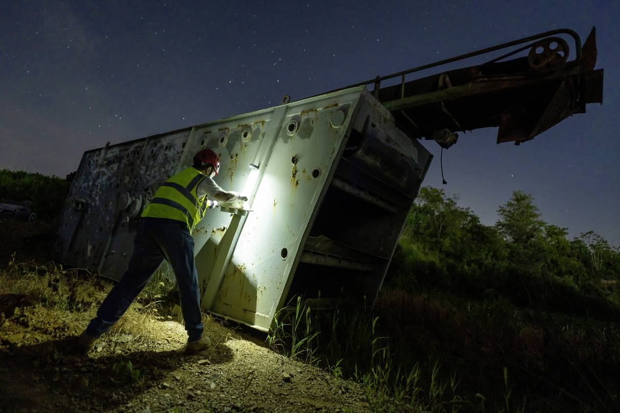 A person wearing a hard hat and hi-vis vest illuminates an old, toppled machine with a torch at night. Stars shine in the sky.