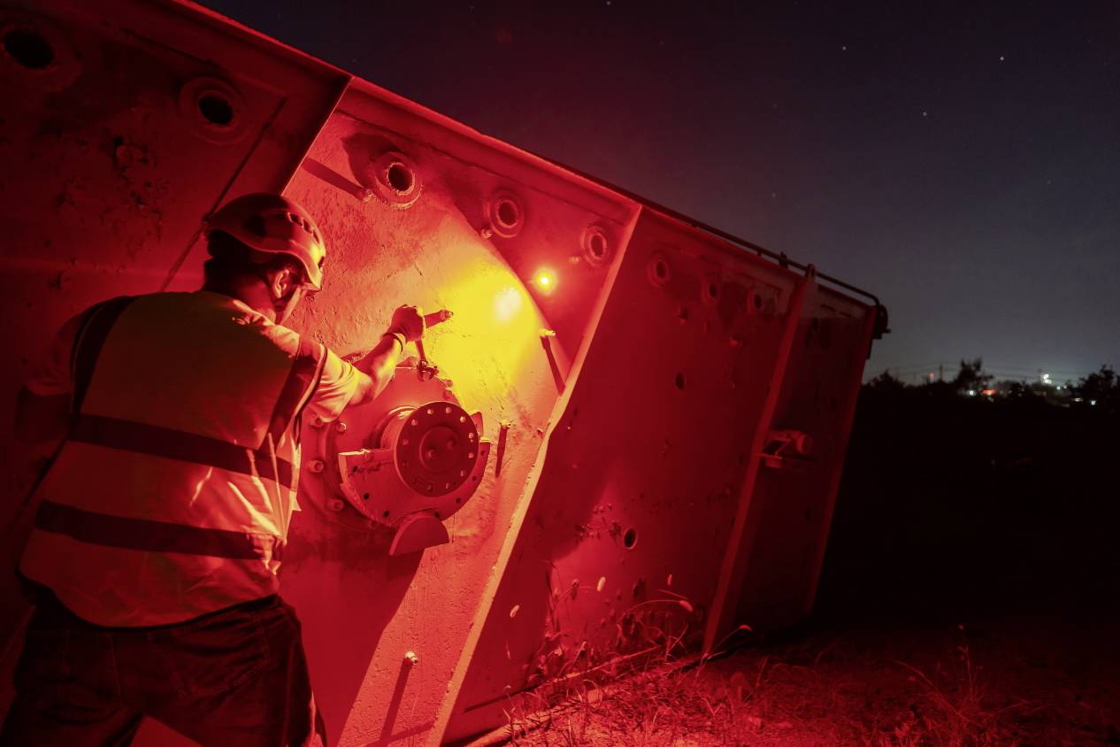 A construction worker wearing a hard hat and high-visibility vest is working at night on a large metal structure bathed in red light.