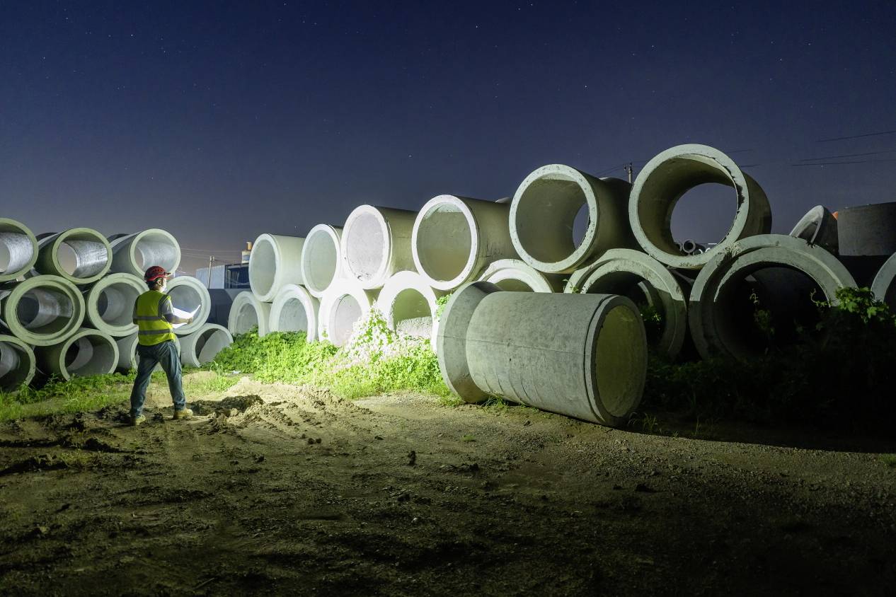 A worker in safety clothing and a hard hat inspects large concrete pipes on a construction site storage area at night, illuminated by a torch.