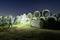 A worker in safety clothing and a hard hat inspects large concrete pipes on a construction site storage area at night, illuminated by a torch.