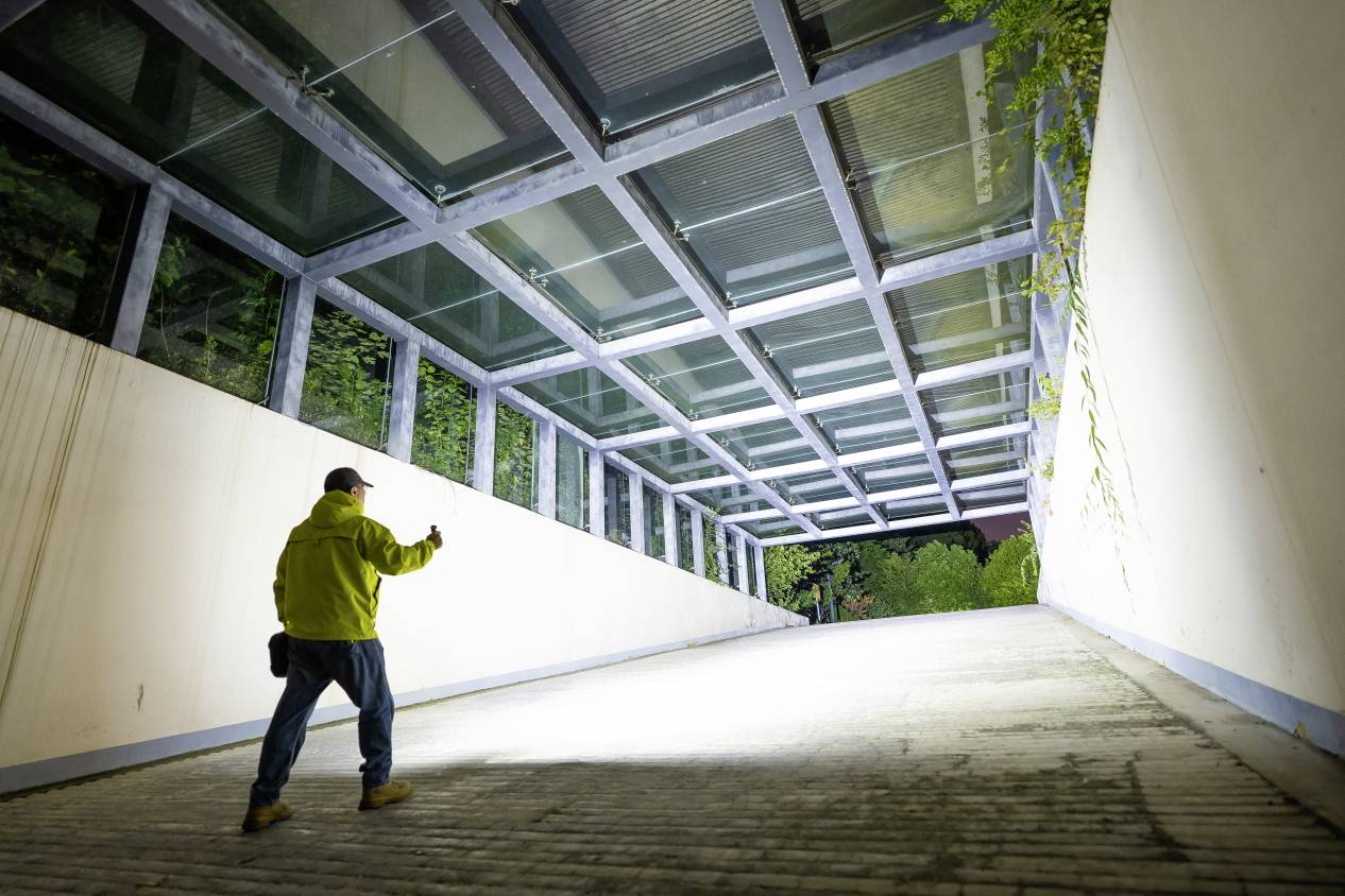 A person wearing light-coloured clothing is photographing an illuminated pathway, surrounded by glass and plants, in a modern setting.