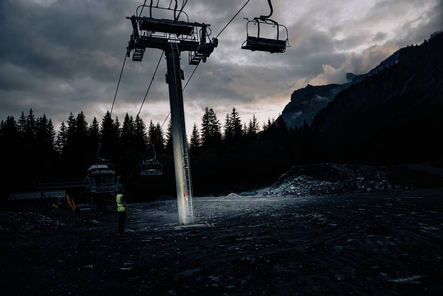 A luminous ski lift surrounded by a mountain landscape at twilight, with a person in reflective clothing looking upwards beneath it.